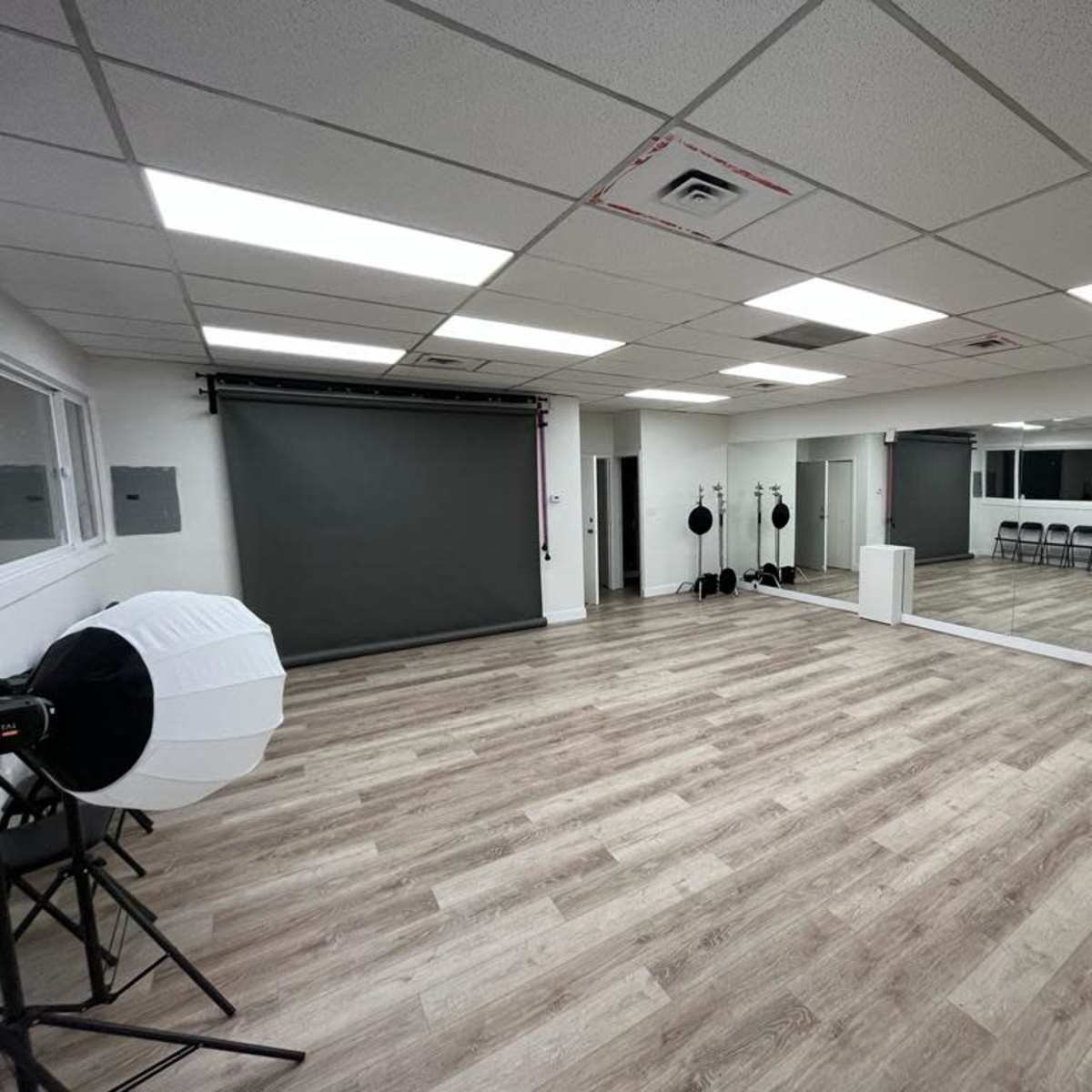 A spacious room with a mirrored wall, rolled-up backdrop, and photography lighting equipment arranged beside rows of folding chairs on a wooden floor.