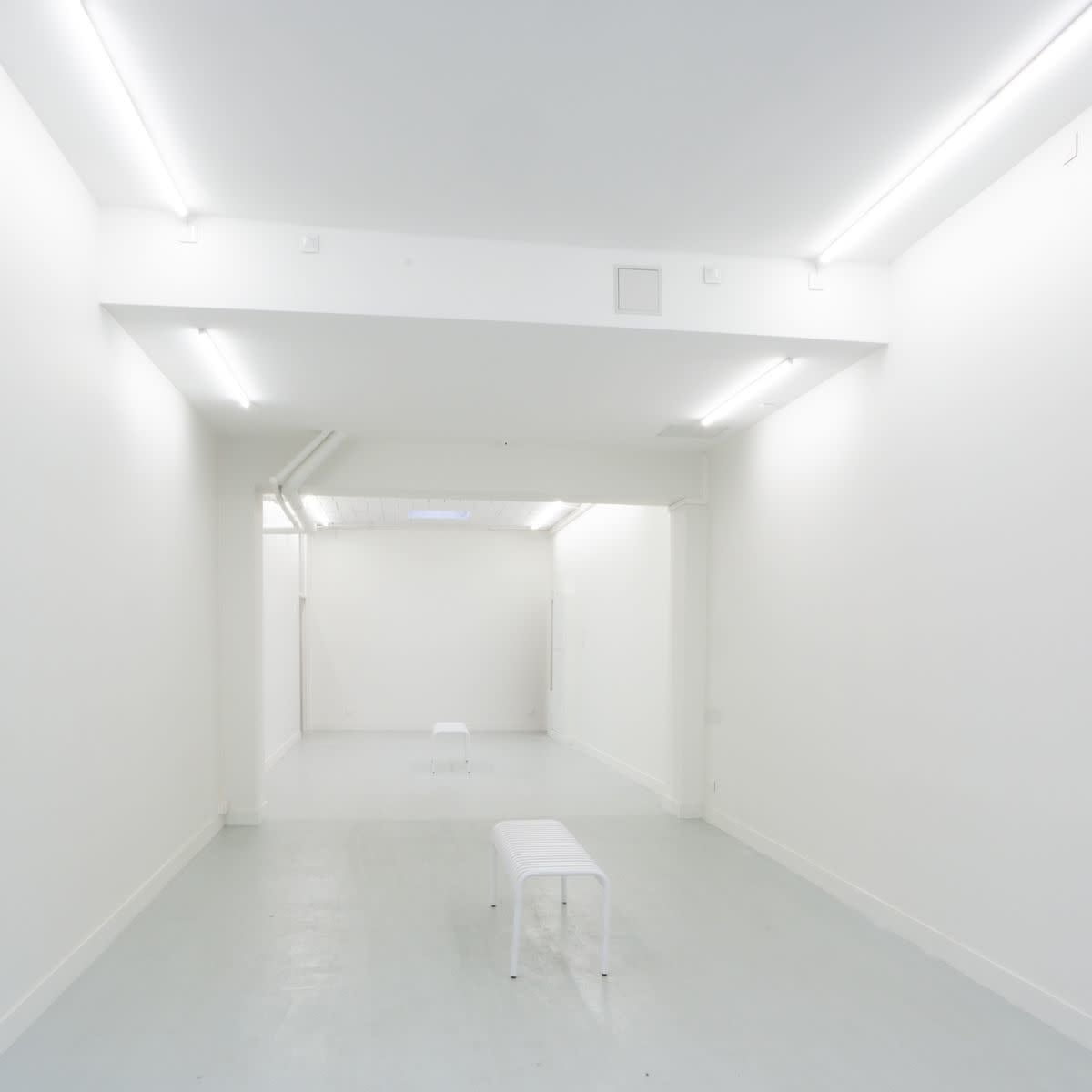 A minimalistic, brightly lit white room featuring two white benches positioned on a light-colored floor.
