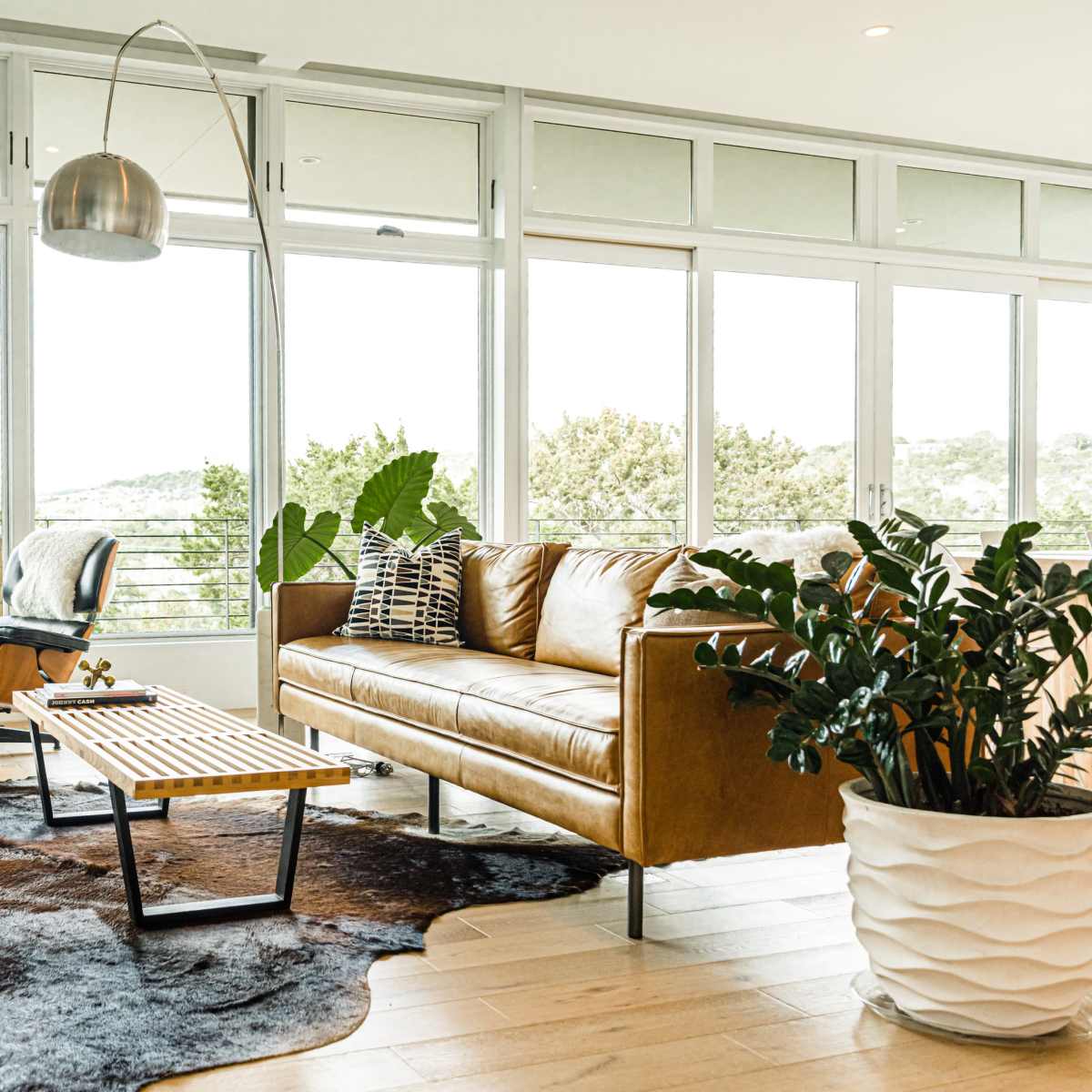 A modern living room features a brown leather sofa, a wooden coffee table, and large windows that offer a view of greenery outside.