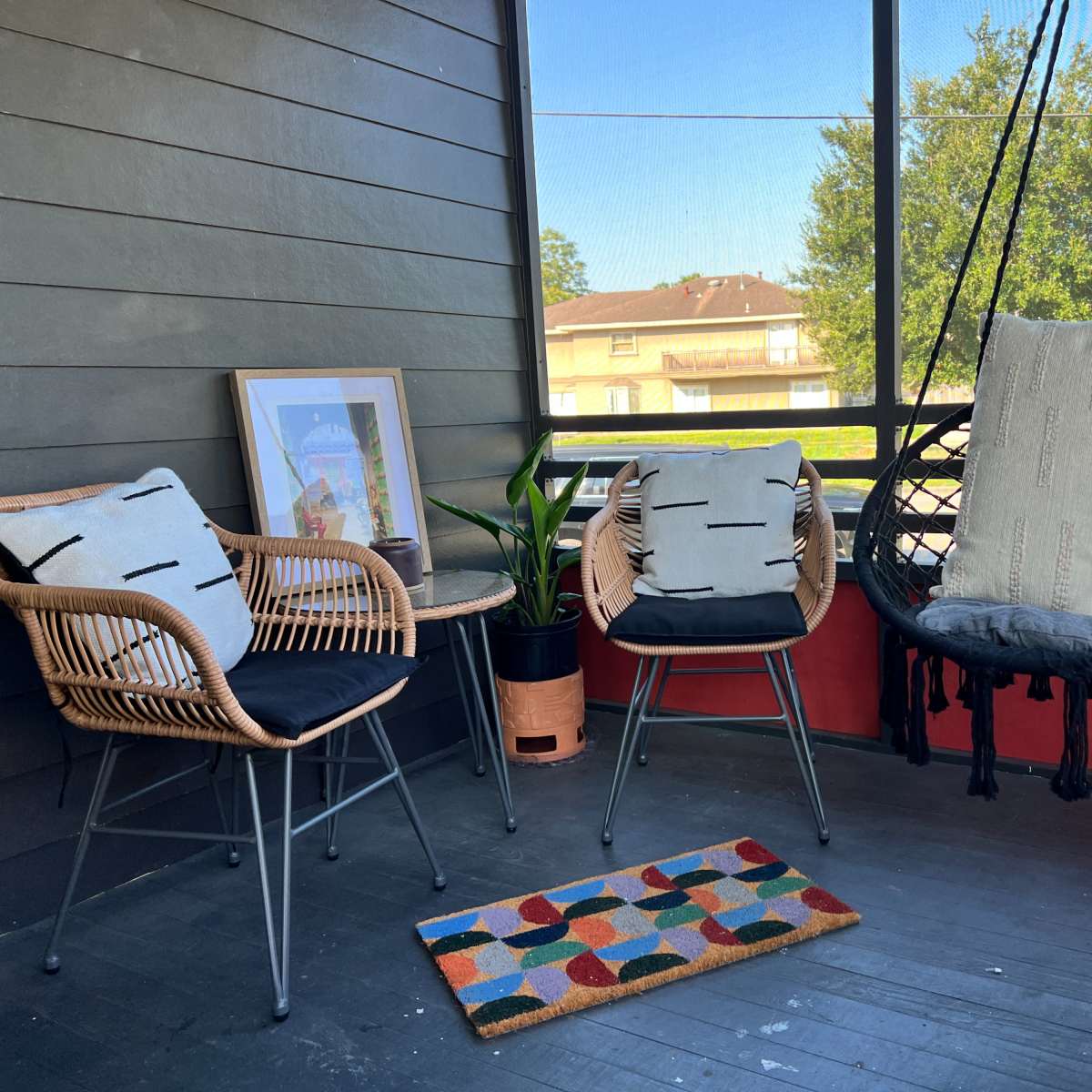 A screened-in porch with two chairs, a hanging chair, a potted plant, and a colorful rug.