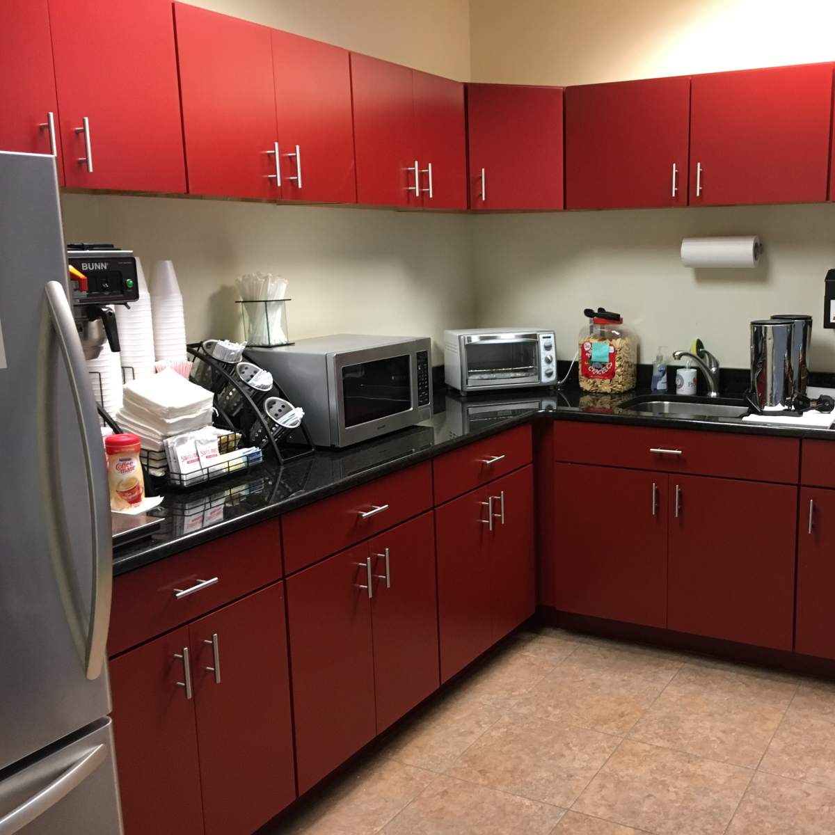 A modern kitchen features red cabinets, a stainless steel refrigerator, microwave, and countertop appliances, with a snack rack displaying various snacks along one wall.