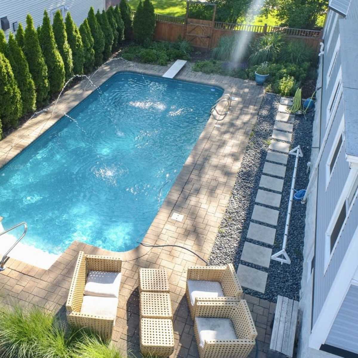 A rectangular swimming pool surrounded by neatly arranged paver stones and a seating area with wicker furniture, lined by tall green shrubs.