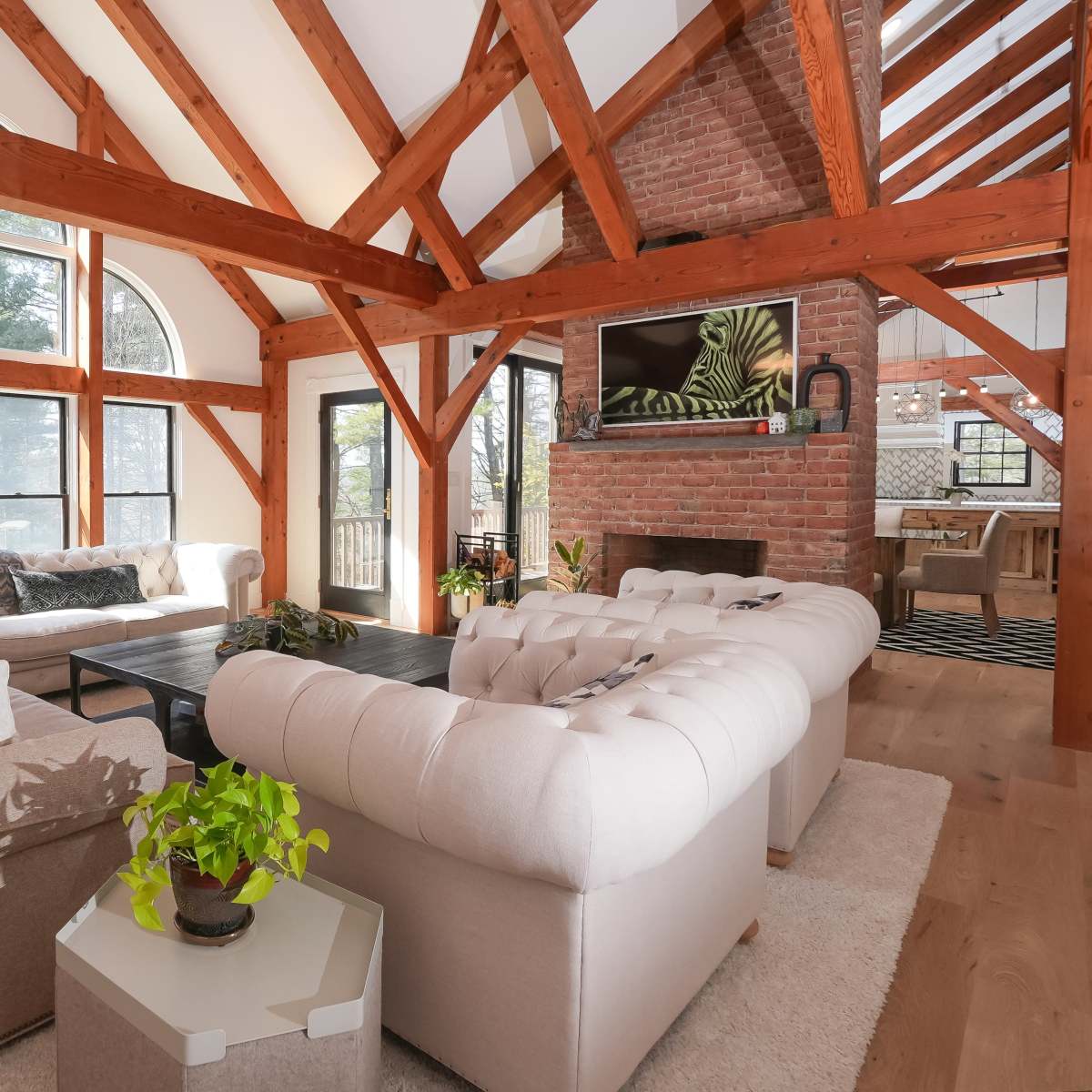 A spacious living room features large windows, exposed wooden beams, and a combination of light-colored sofas arranged around a coffee table.