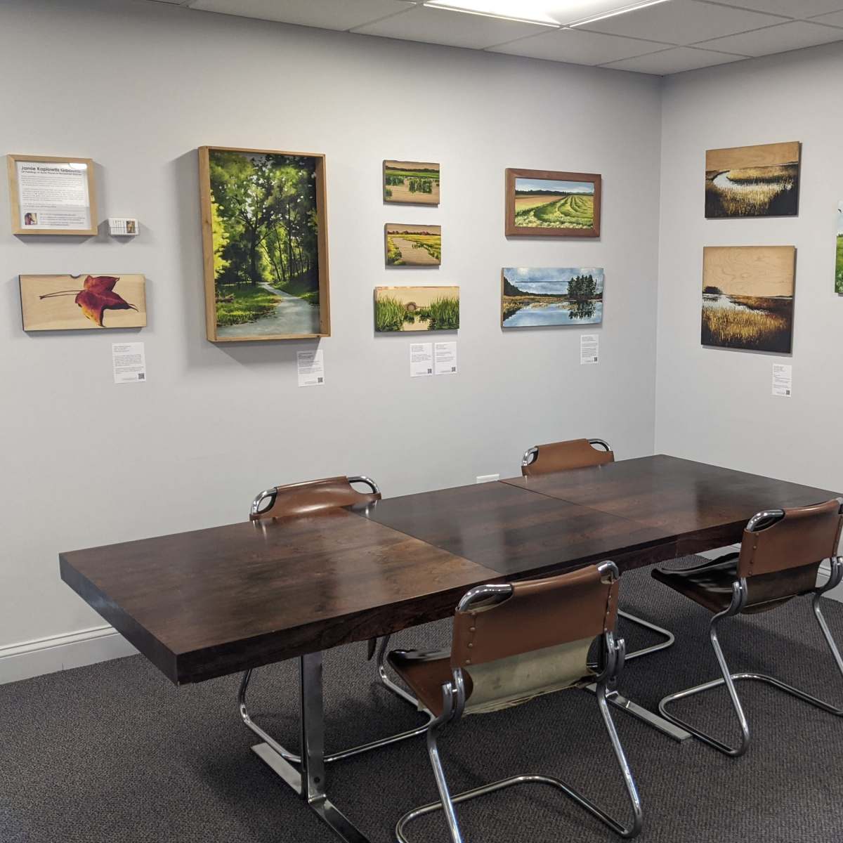 A conference room with a large wooden table surrounded by four brown chairs, and walls adorned with various landscape paintings and informational plaques.
