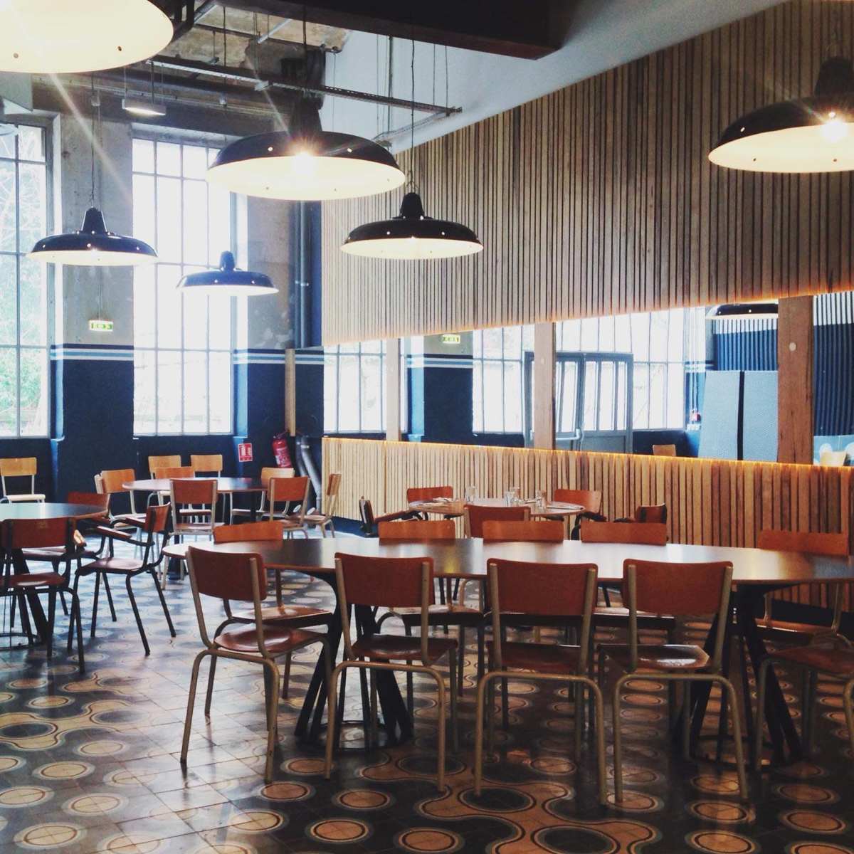 A spacious, modern café interior with several wooden tables and chairs, large windows allowing natural light, and industrial-style lighting fixtures.