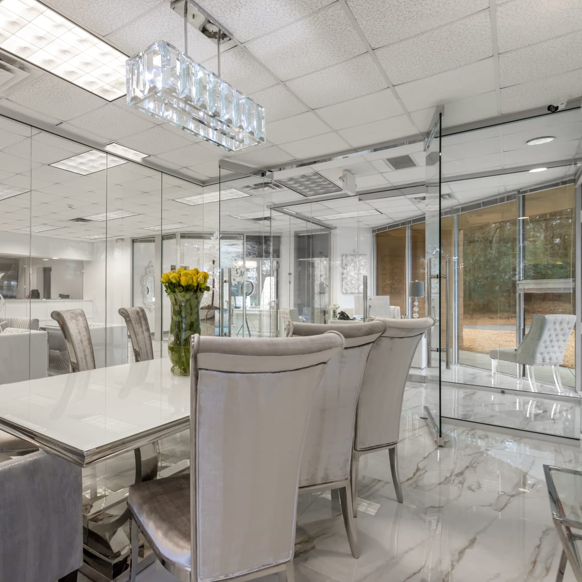A modern conference room with a glass-enclosed design, featuring a white table and plush, light gray chairs arranged around it, with a floral centerpiece and ample lighting from the ceiling.