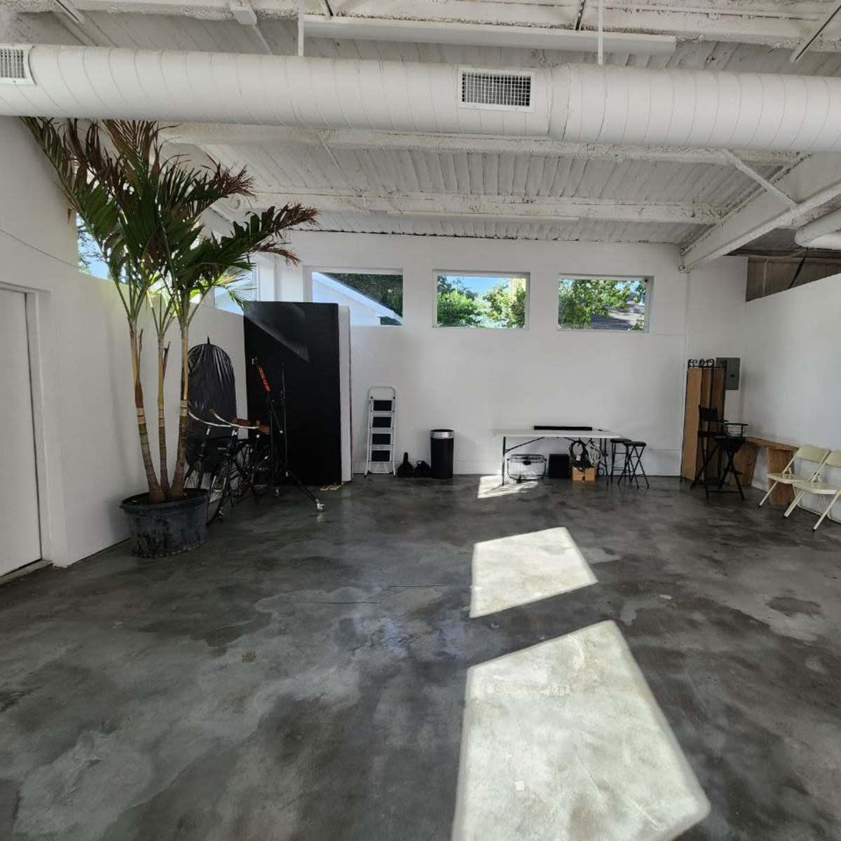 A spacious, minimally furnished room with a concrete floor, a tall palm plant in a pot, and various tables and chairs arranged along the walls.