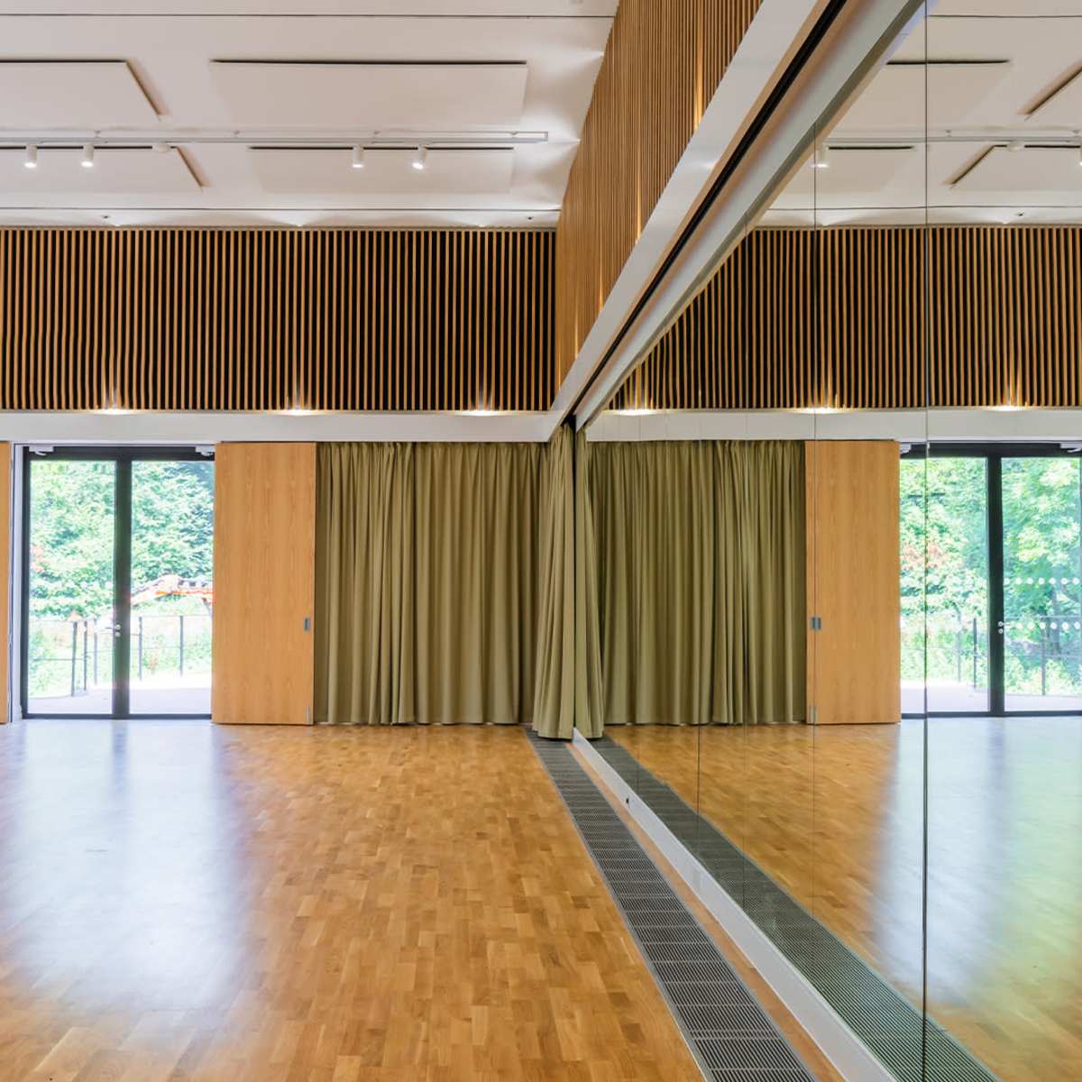 A spacious, wooden-floored room with large mirrors on one wall and floor-to-ceiling curtains on either side of a pair of glass doors leading to an outdoor area.
