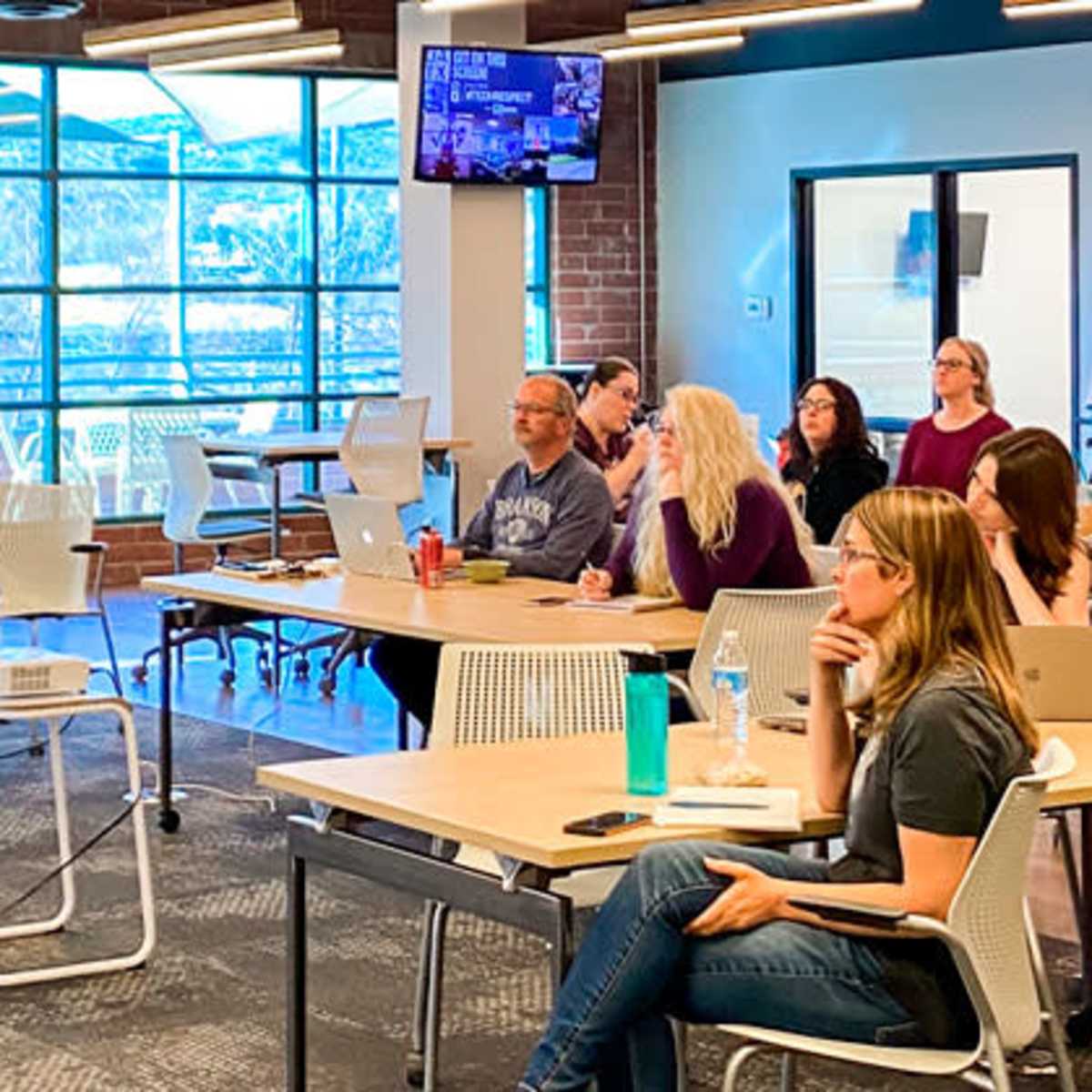 A presenter stands in front of a group of seated attendees in a modern conference room with large windows and a presentation screen.