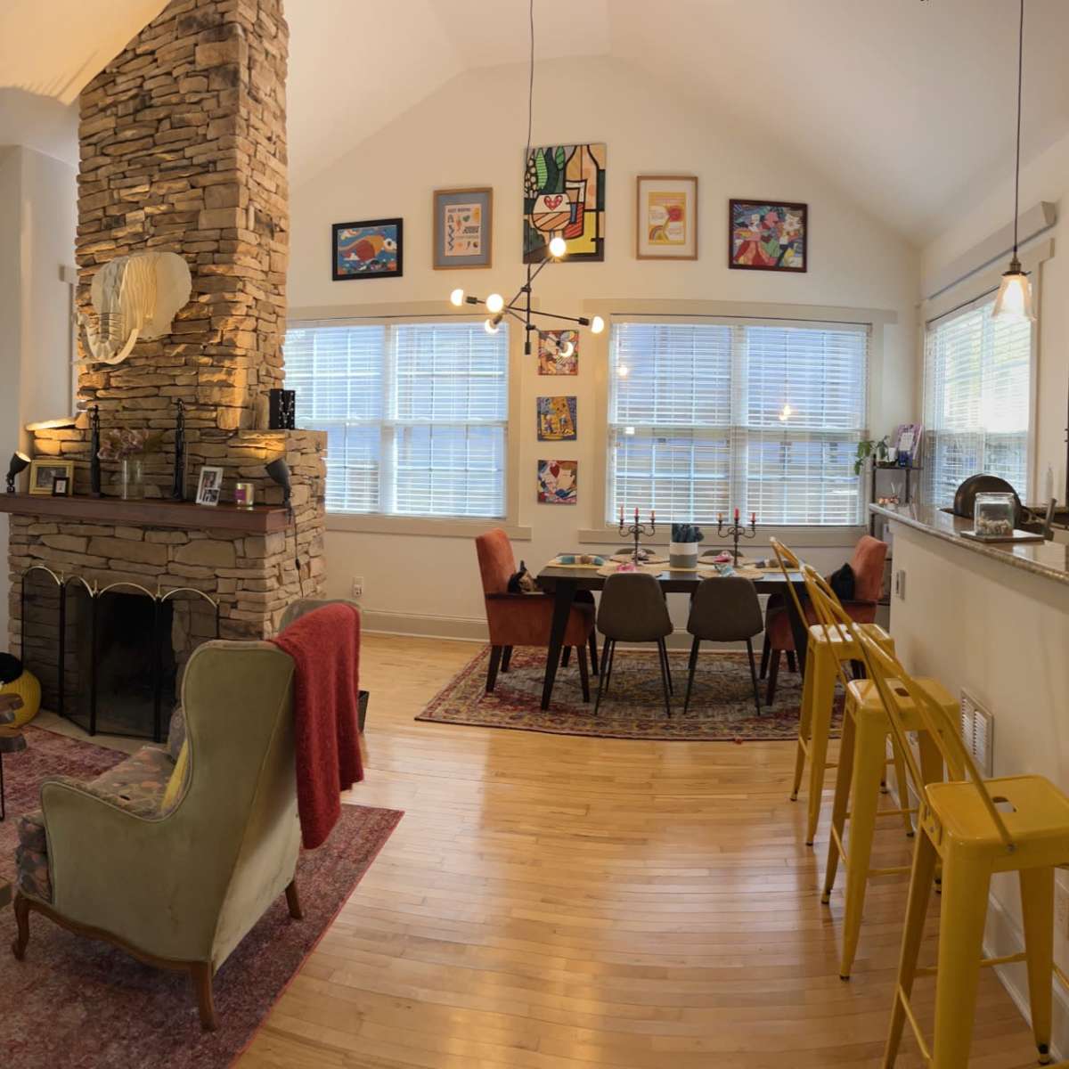An open-concept living space featuring a living area with colorful chairs, a stone fireplace, a dining table, and a kitchen with wooden cabinets.