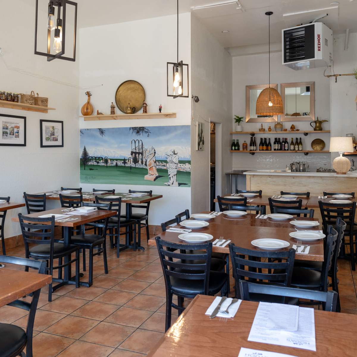 The interior of a restaurant features wooden tables and chairs, art on the walls, and a serving area with a countertop and shelves.