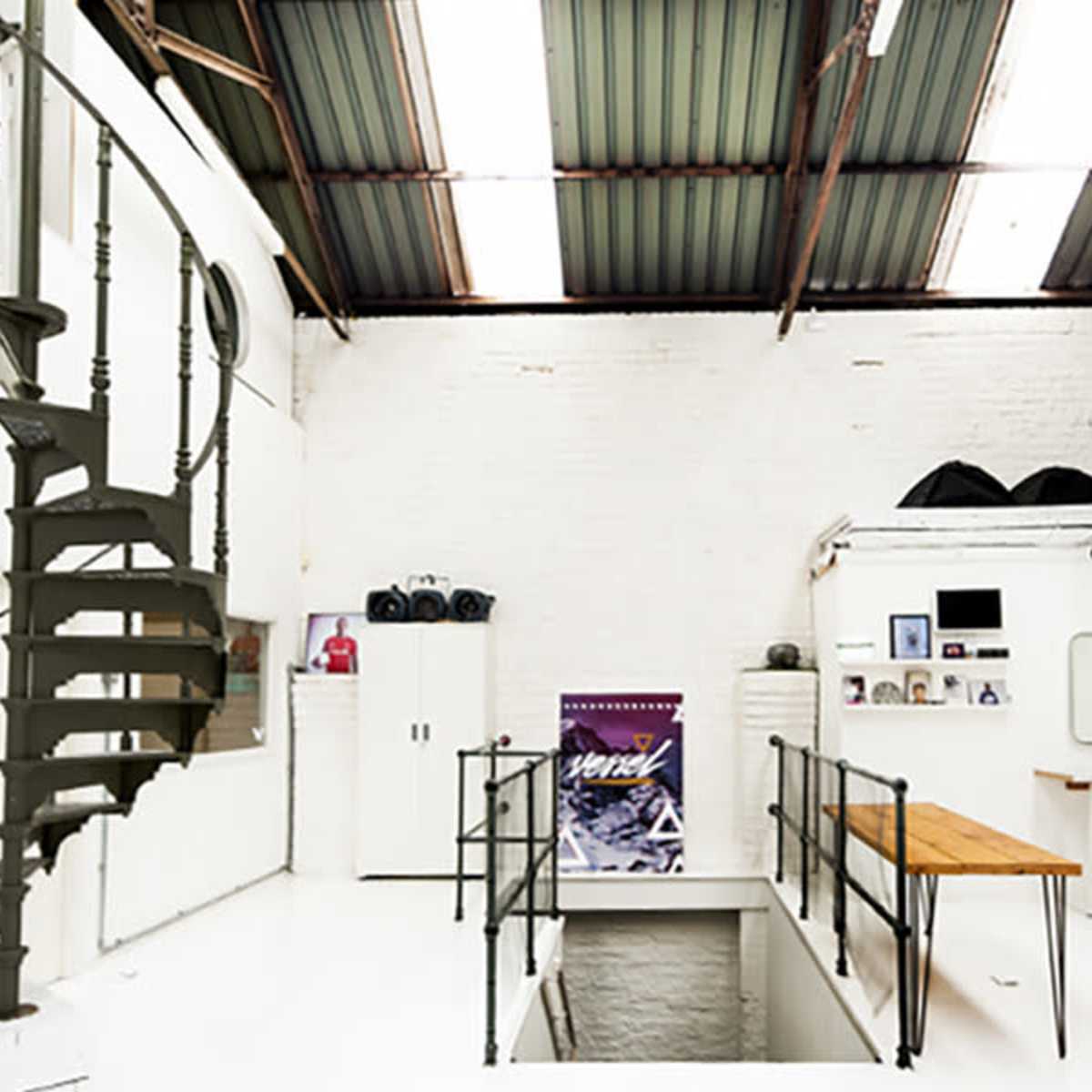 A modern, industrial-style studio with a spiral staircase, white walls, and various furniture pieces, including barstools and a long wooden table.