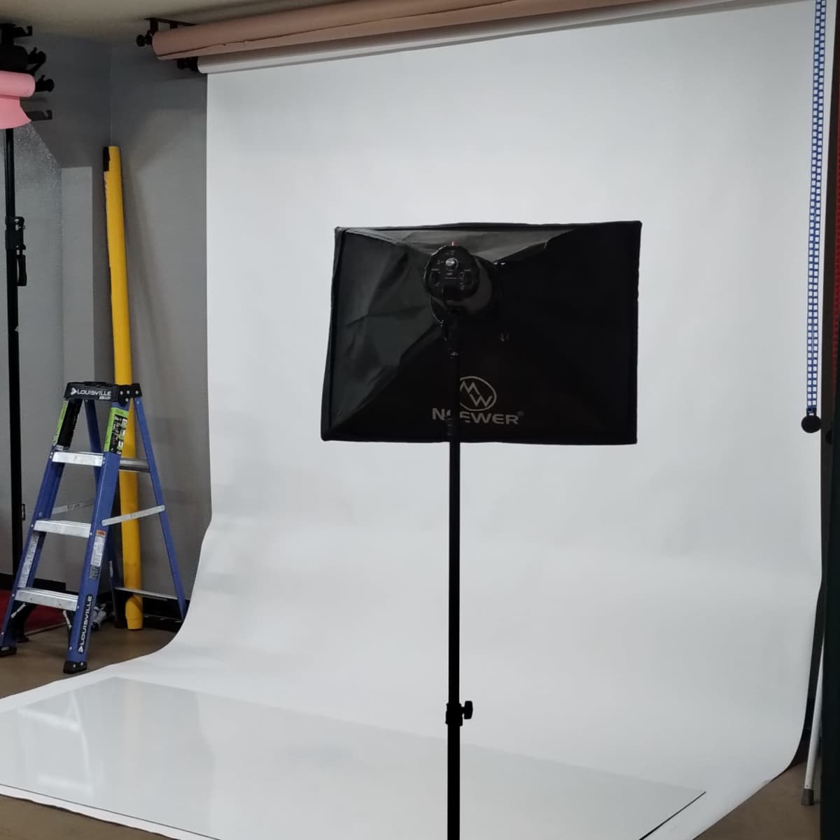 A photography studio setup with two light stands, a white backdrop, and a ladder in the background.