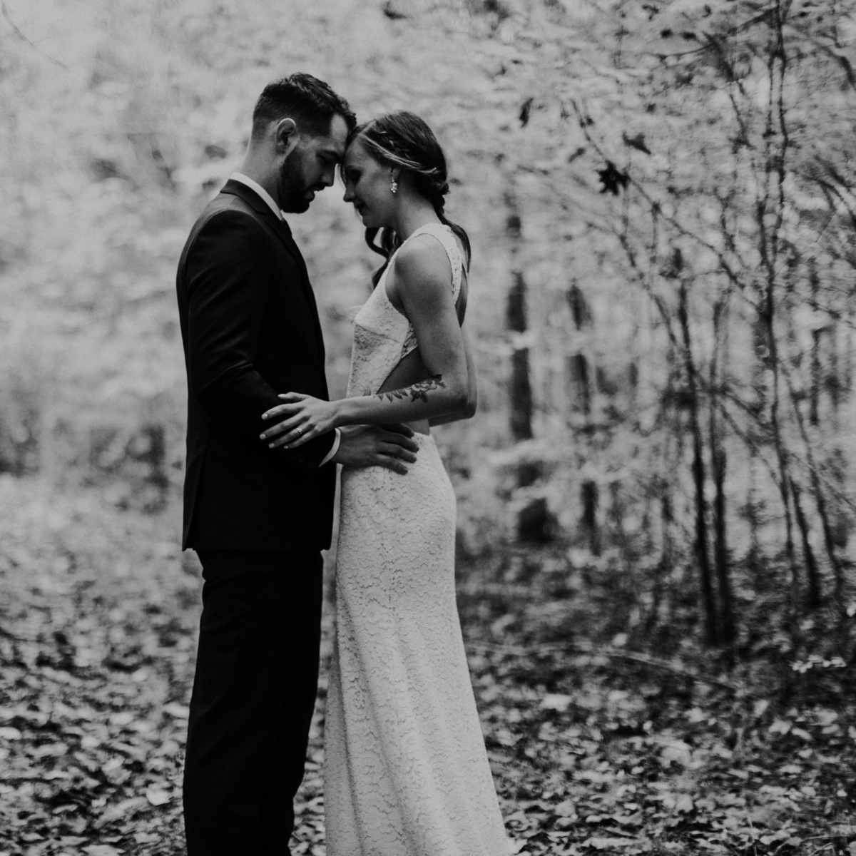 A couple stands close together in a forest, surrounded by trees and foliage.