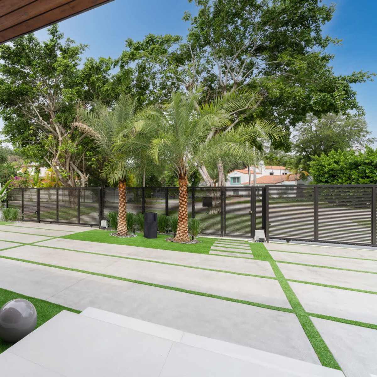A modern outdoor patio features a landscaped area with palm trees, green grass, and decorative stone elements surrounded by a black fence.