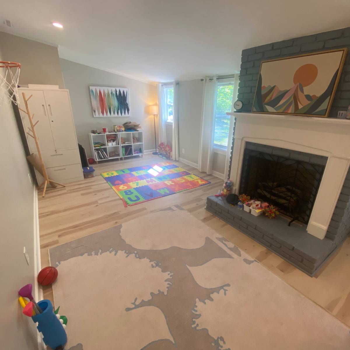 A playroom features a colorful rug with letters, a basketball hoop, a fireplace with a gray brick facade, and a window allowing natural light.