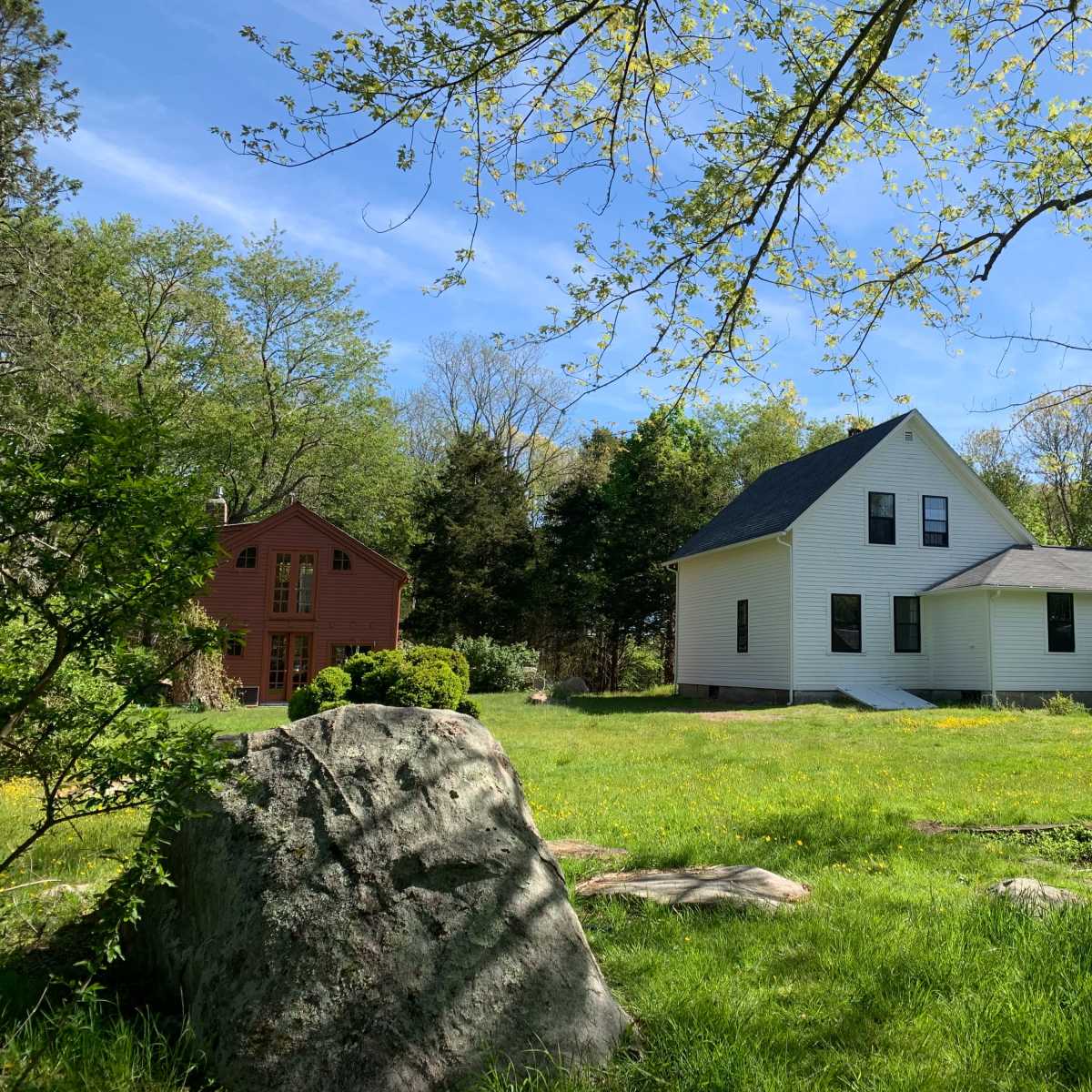Two houses, one white and one red, situated on a grassy area surrounded by trees and large rocks.