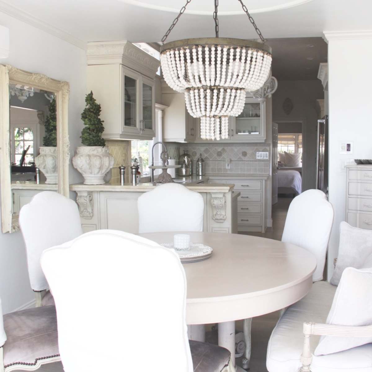 A dining area features a round table surrounded by upholstered chairs and a beaded chandelier, with a kitchen visible in the background.