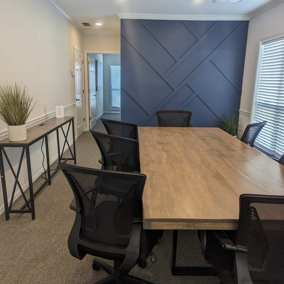 A conference room with a wooden table surrounded by black mesh chairs, featuring a blue accent wall and potted plants.