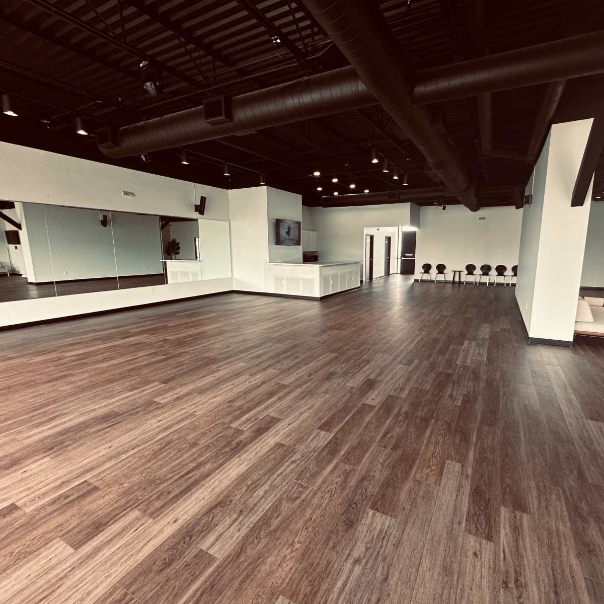 A spacious, empty studio with wooden flooring, large mirrors on one wall, and sleek furniture arranged throughout.