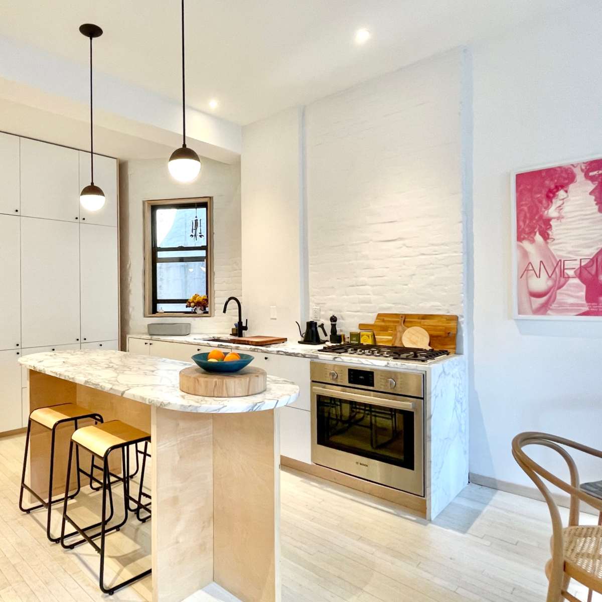 A modern kitchen features a marble island with bar stools, stainless steel appliances, and white cabinetry, illuminated by pendant lights.