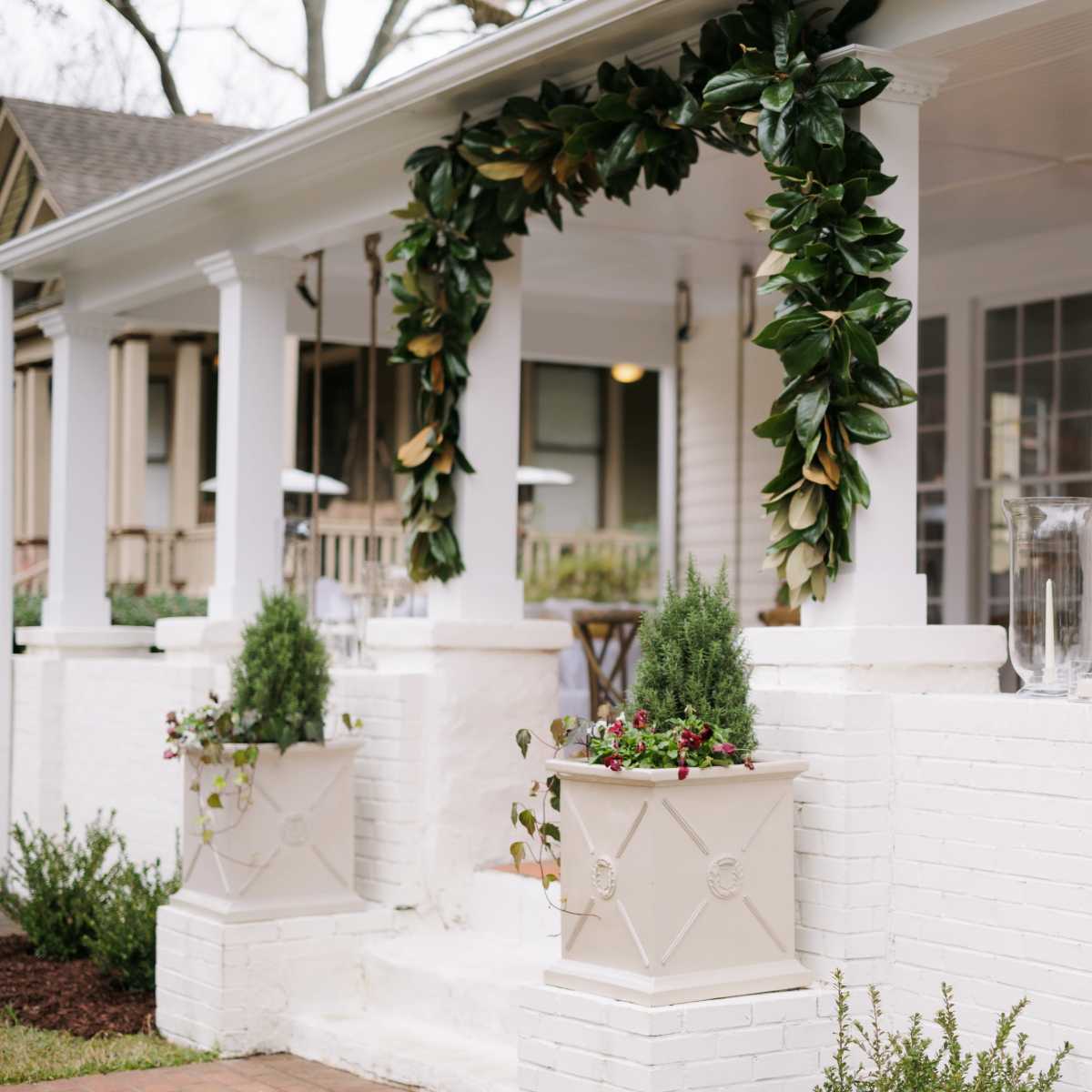 The porch features white columns adorned with a green garland and potted plants alongside decorative glass containers on a white wall.