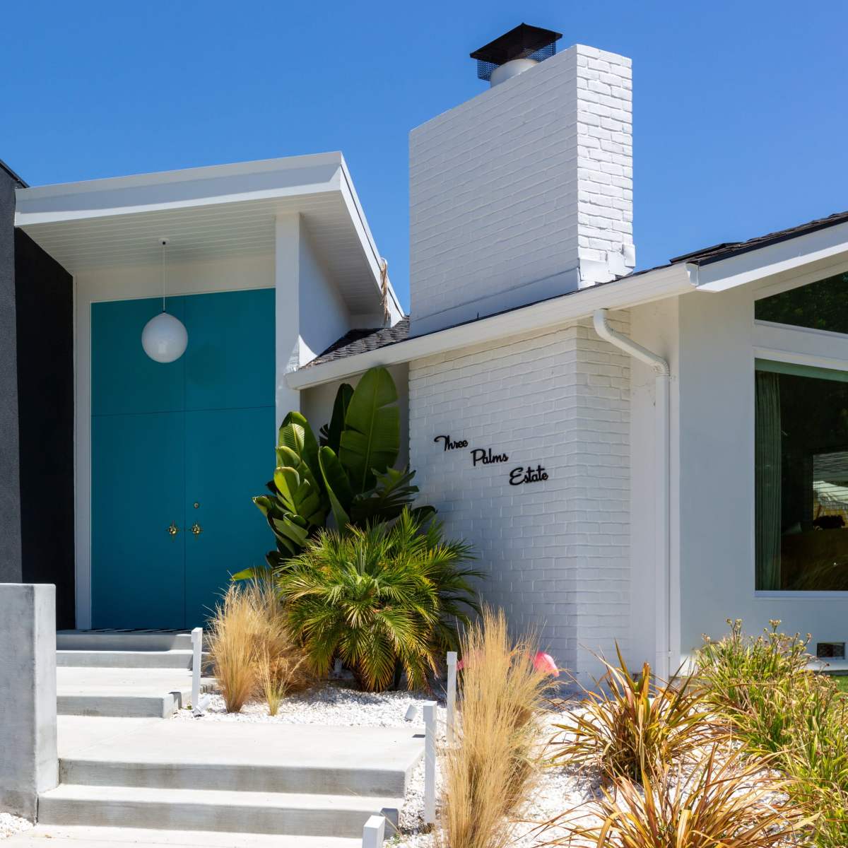 A modern house features a contrasting black wall, a bright turquoise door, and a landscaped front yard with various plants.