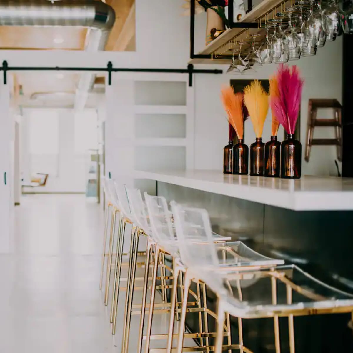 A modern bar area with clear stools and colorful decorative vases on the counter, flanked by sliding doors leading to a bright space.