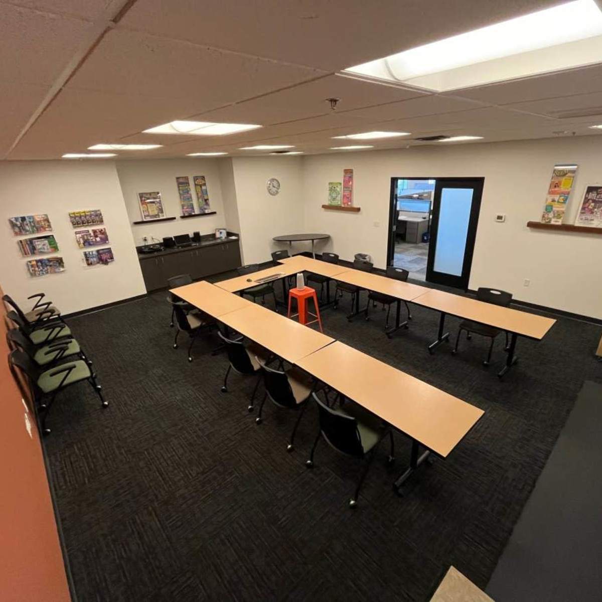 A meeting room with several tables arranged in a U-shape, black chairs, and a single orange stool in the center.