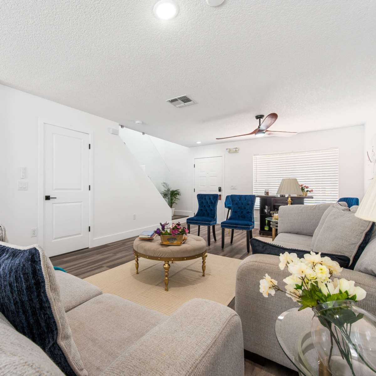 A modern living room with neutral-colored sofas, a round coffee table, and blue upholstered chairs, featuring a ceiling fan and natural light from a window.