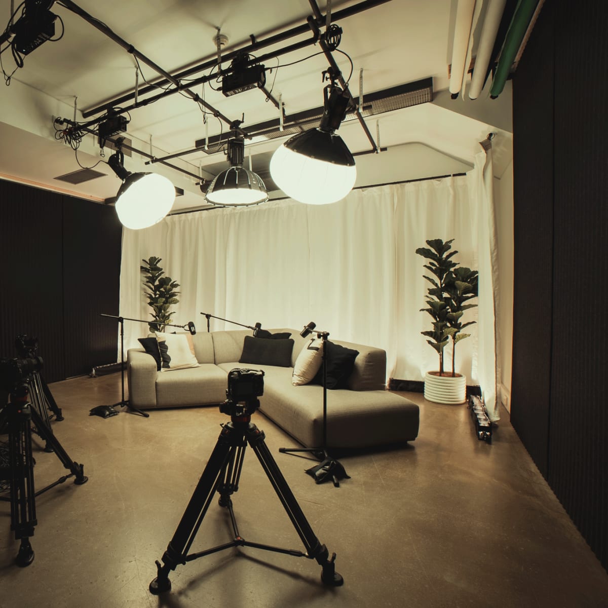 A multimedia studio is set up with two sofas, a white backdrop, and three cameras on tripods.