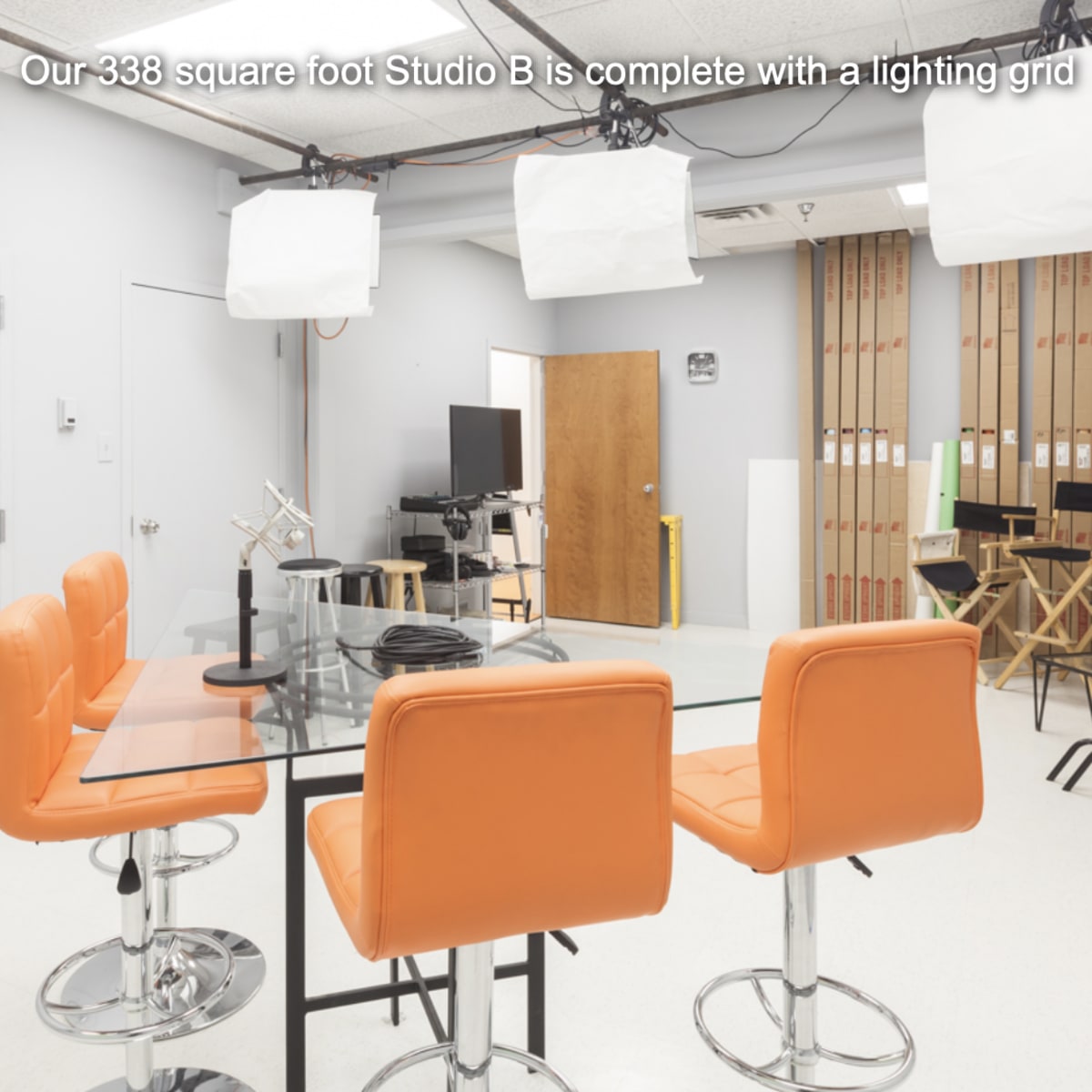 A well-lit studio with orange bar stools around a glass table, equipped with a lighting grid and various studio equipment in the background.