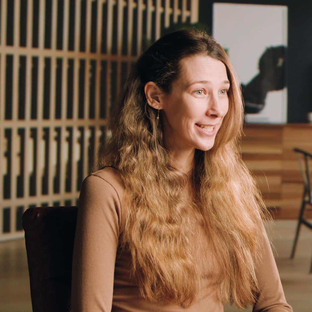 A woman with long, wavy hair sits in a modern office space with wooden furniture and large windows in the background.