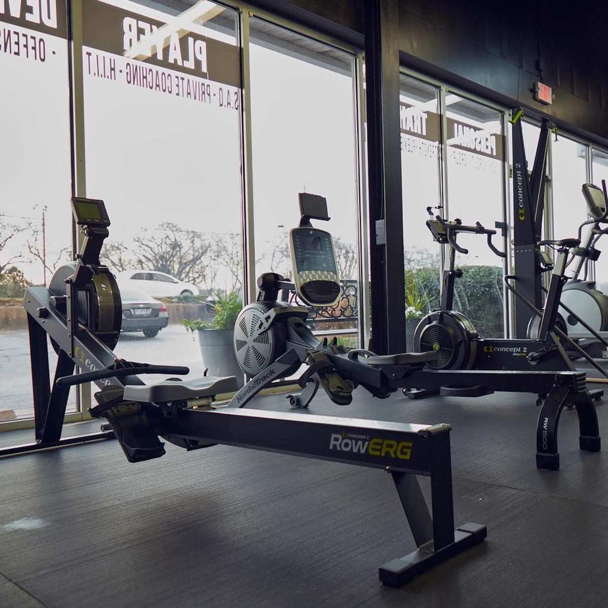 A fitness facility with rowing machines, stationary bikes, and elliptical machines arranged against large windows.