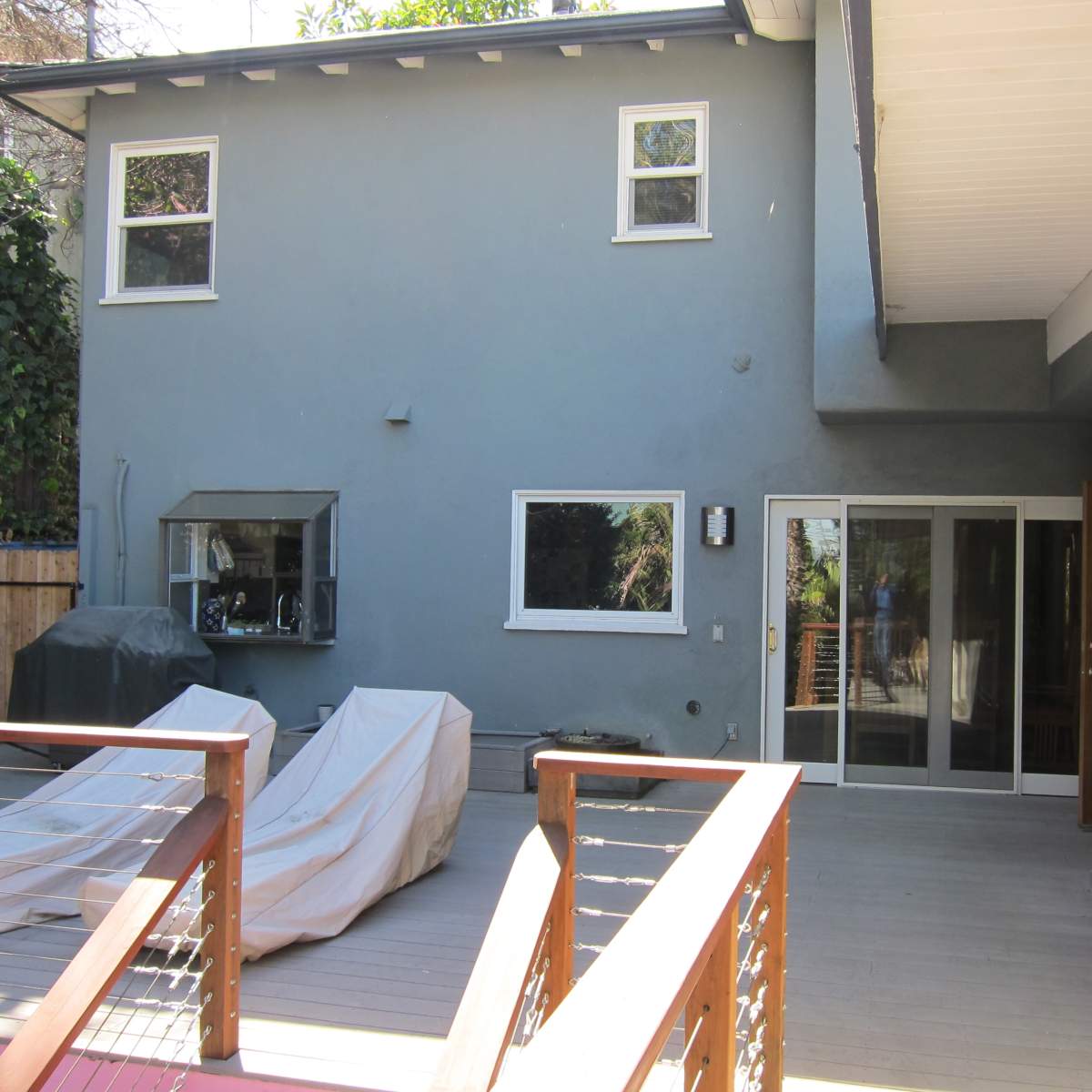 A modern house exterior featuring a wooden deck, covered furniture, and large glass doors leading into the interior.