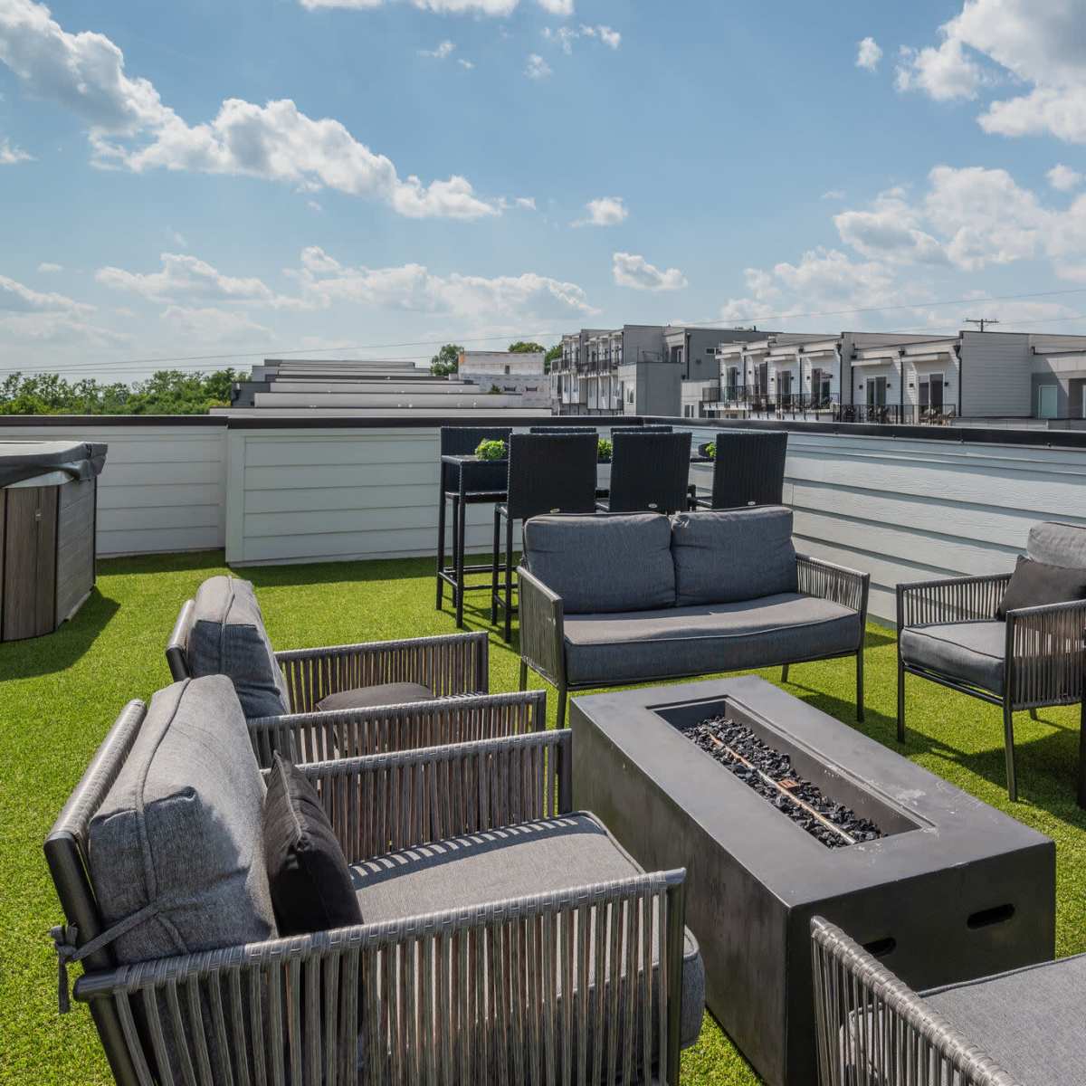 A rooftop terrace features gray furniture arranged around a fire pit, with green synthetic grass and a backdrop of buildings under a partly cloudy sky.