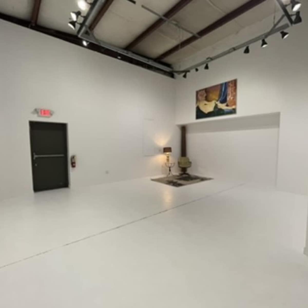 An empty, well-lit room with white walls, a wooden ceiling, a single chair on a rug, and a piece of art hanging on the wall.