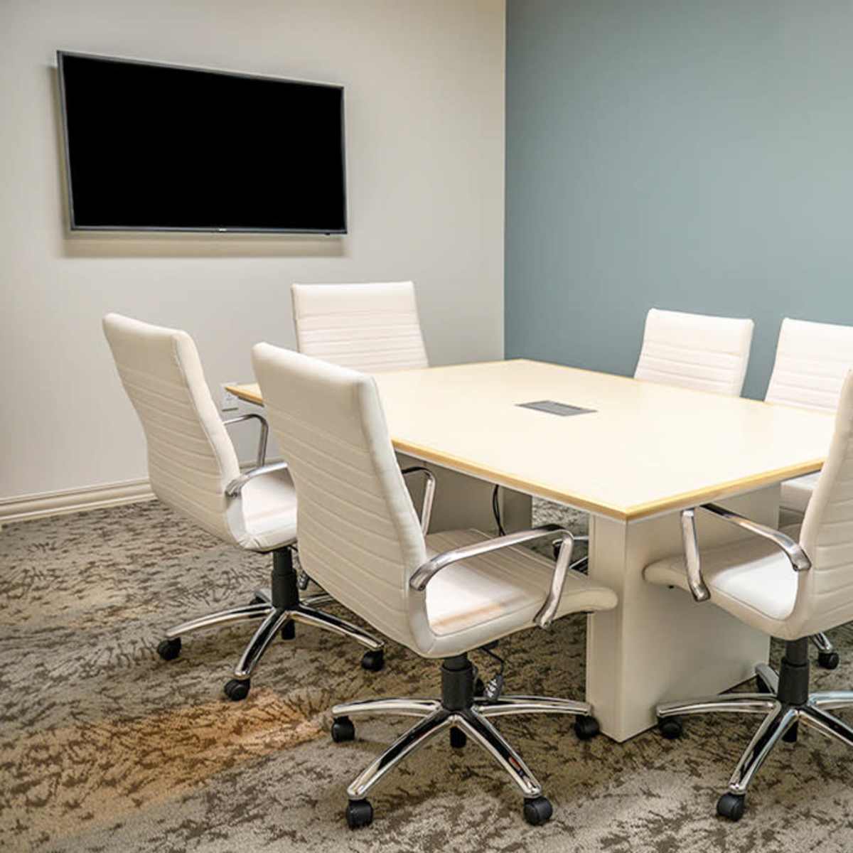 A conference room features a rectangular table surrounded by five white chairs and a wall-mounted screen.
