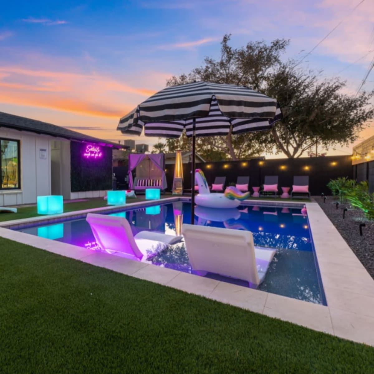 A modern backyard with a swimming pool, colorful illuminated furniture, and a large striped umbrella set against a sunset sky.