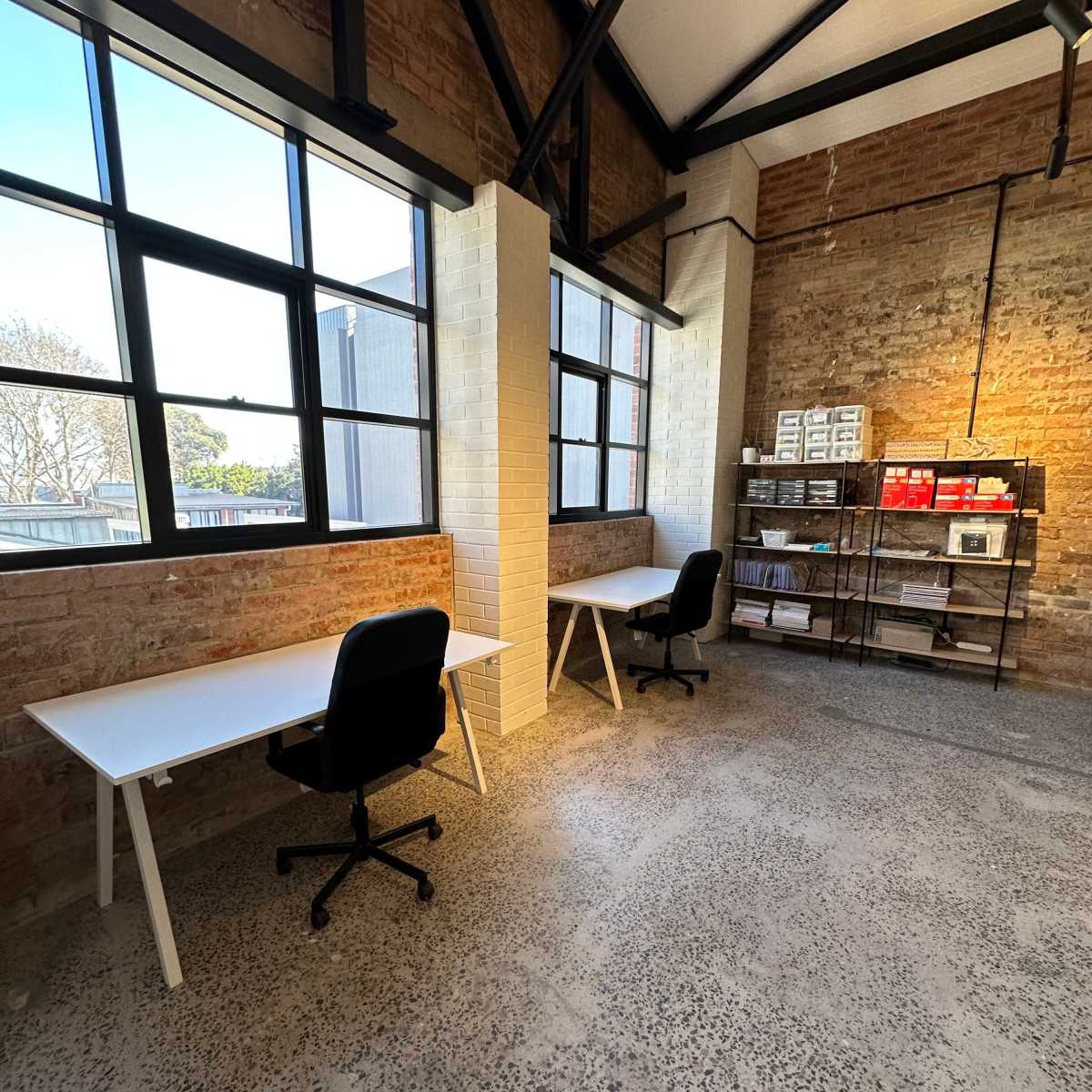 A spacious office area with two white desks and black chairs, illuminated by natural light from large windows, alongside a shelving unit containing various materials against a brick wall.