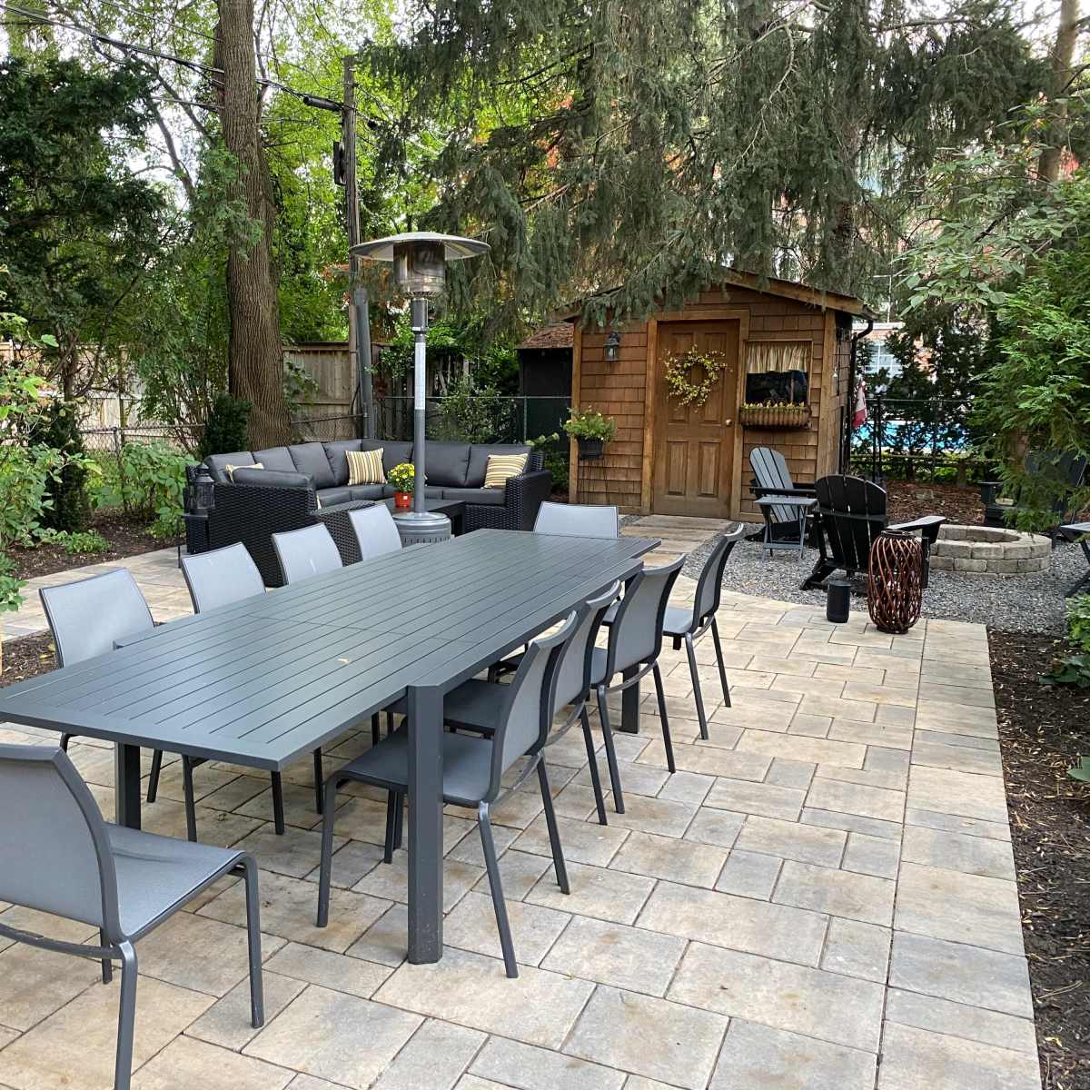 An outdoor patio area with a long dining table surrounded by chairs, a seating area, a patio heater, and a wooden shed in the background.