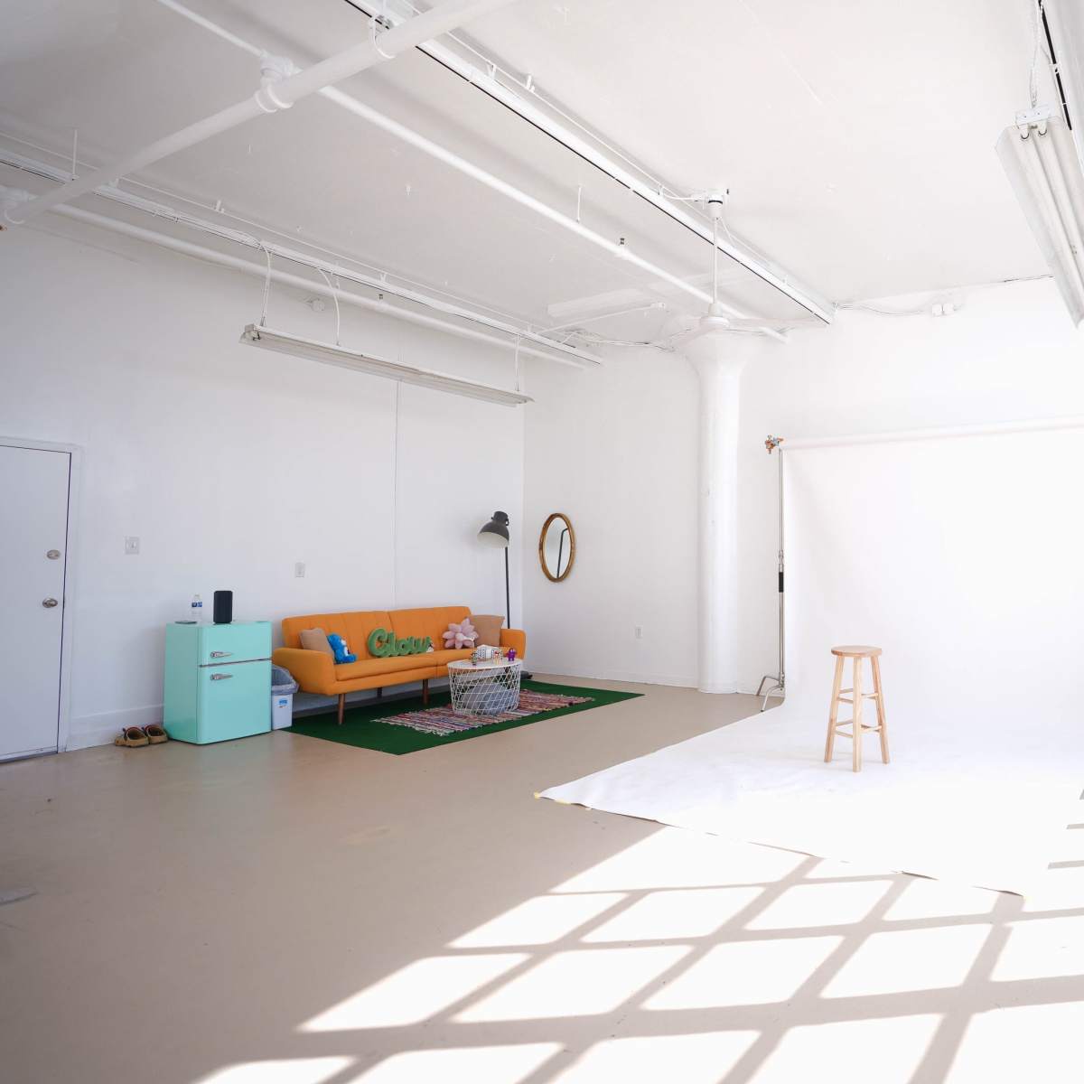 A bright, spacious studio with a white backdrop, a wooden stool in the center, a teal refrigerator, a couch, and large windows letting in natural light.