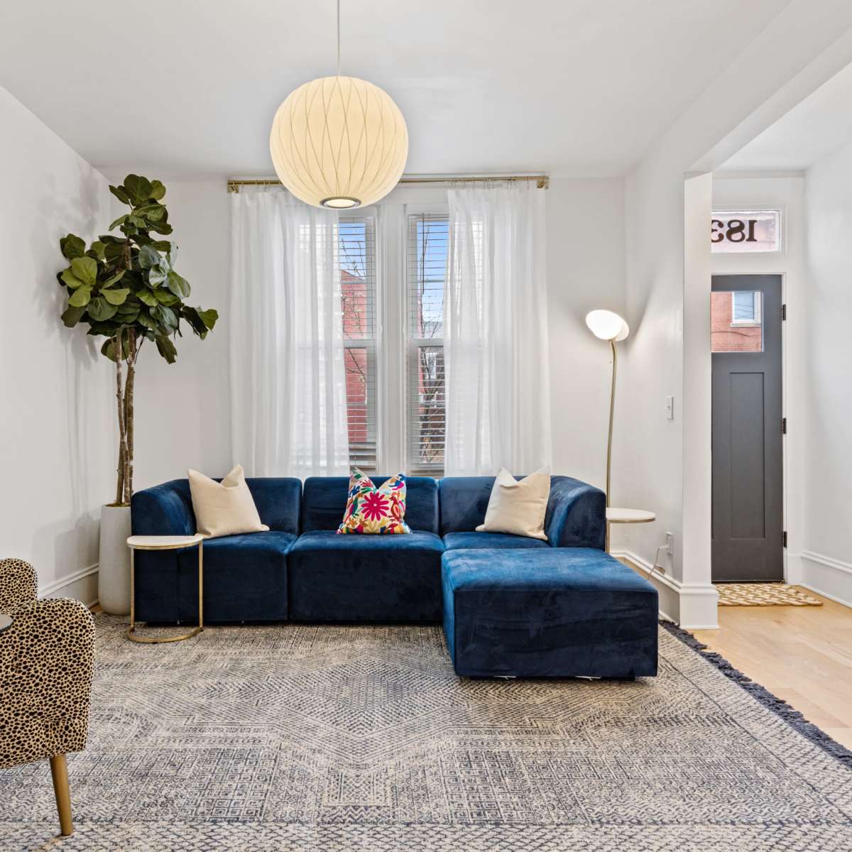 The living room features a blue sectional sofa, a patterned chair, a floor plant, and a round pendant light, complemented by a rug and framed artwork on the walls.