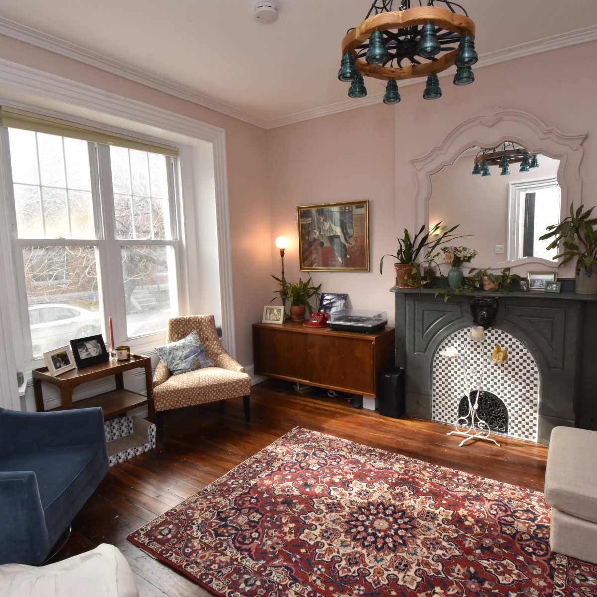 A cozy living room featuring a dark green fireplace, a beige sofa, a blue armchair, and a patterned rug on wooden floors.