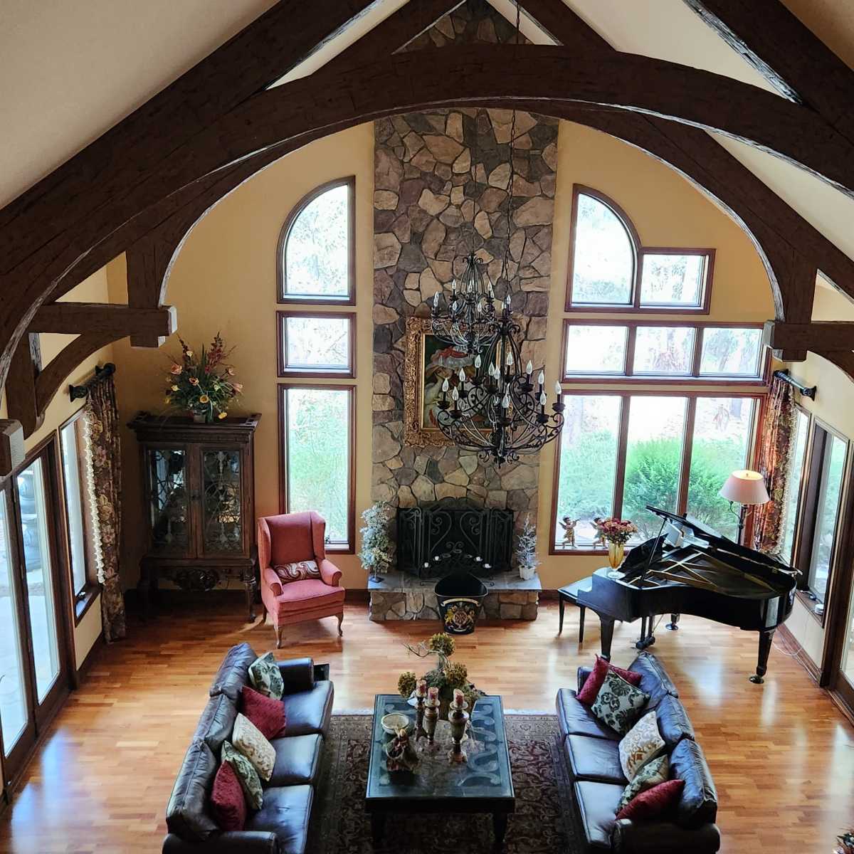 A spacious living room with high ceilings, wooden beams, a grand piano, and large windows overlooking a garden, decorated for the holidays.