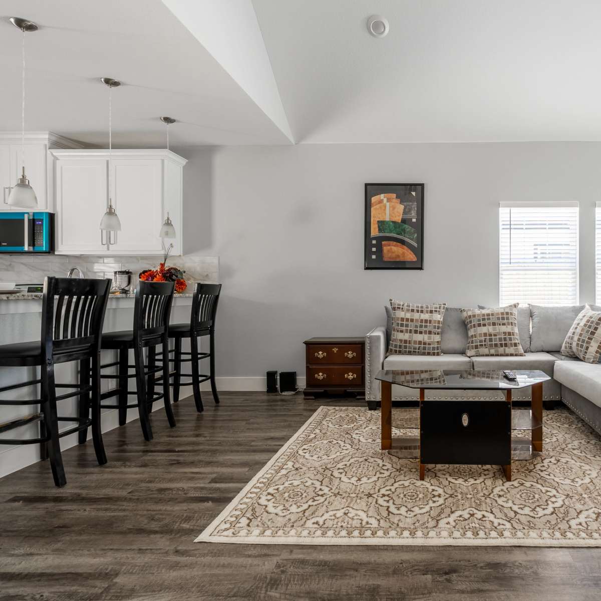 A modern living room with a large sectional sofa, a coffee table, and two framed artworks, adjacent to a kitchen with bar seating.