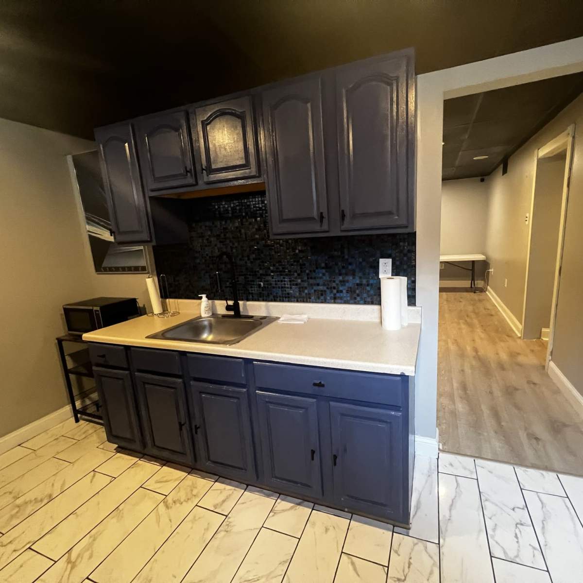 A small kitchen with dark blue cabinets, a sink, and a textured backsplash, leading to a hallway with light wood flooring.