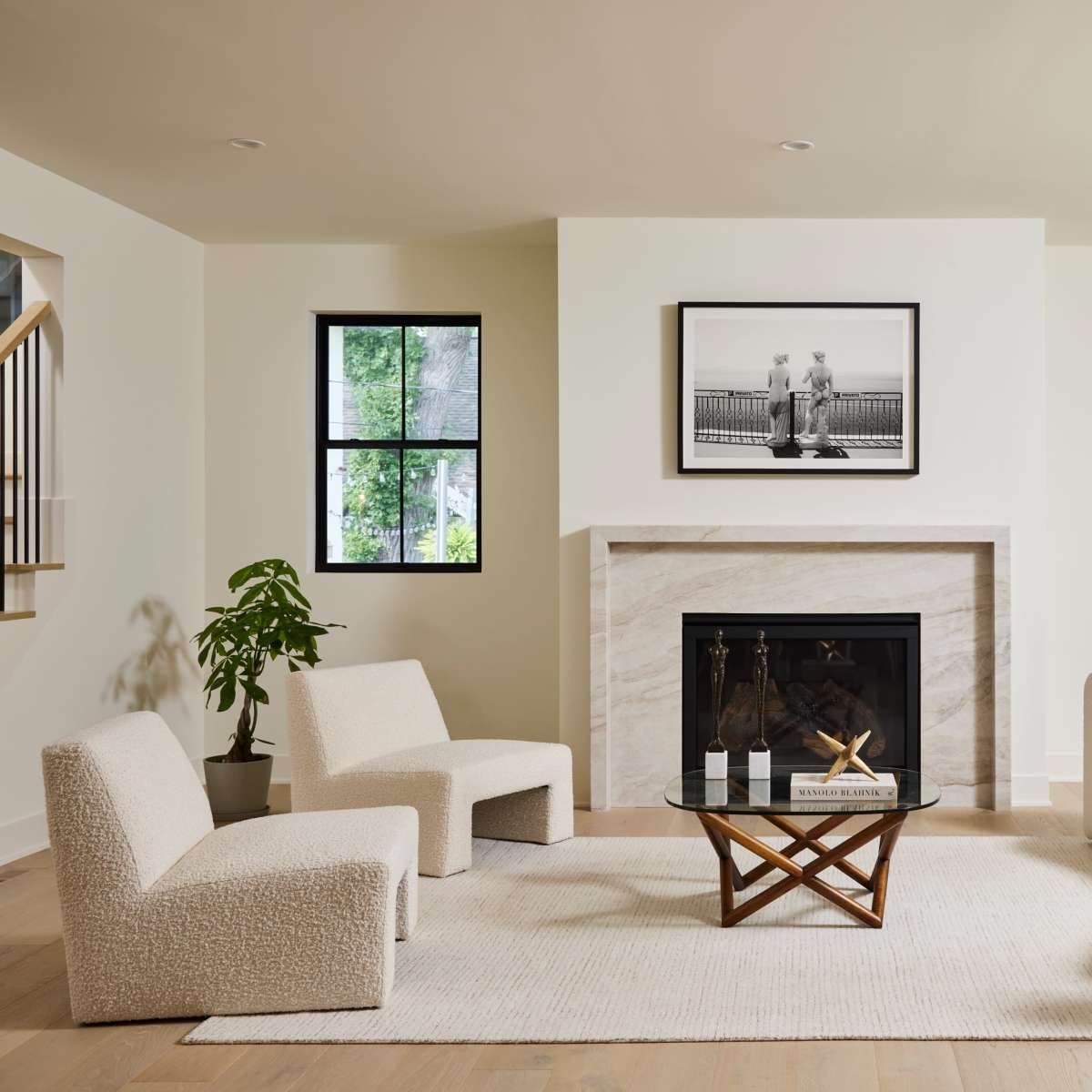 A modern living room features two upholstered chairs, a round sofa, a glass coffee table on a rug, and a fireplace with a framed black-and-white photograph above it.