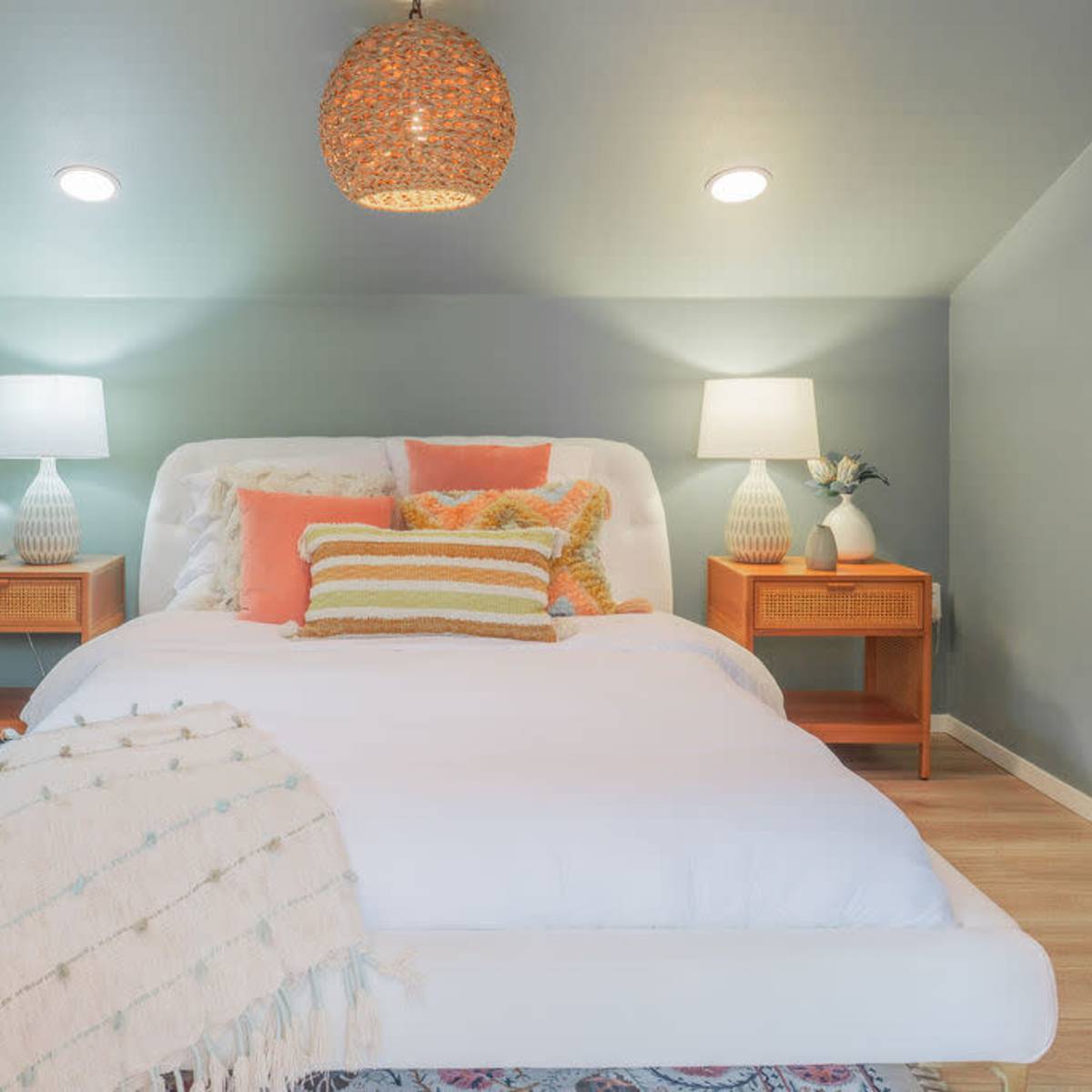 A neatly arranged bedroom features a white bed with colorful pillows, two matching bedside lamps, and textured decor against a soft green wall.