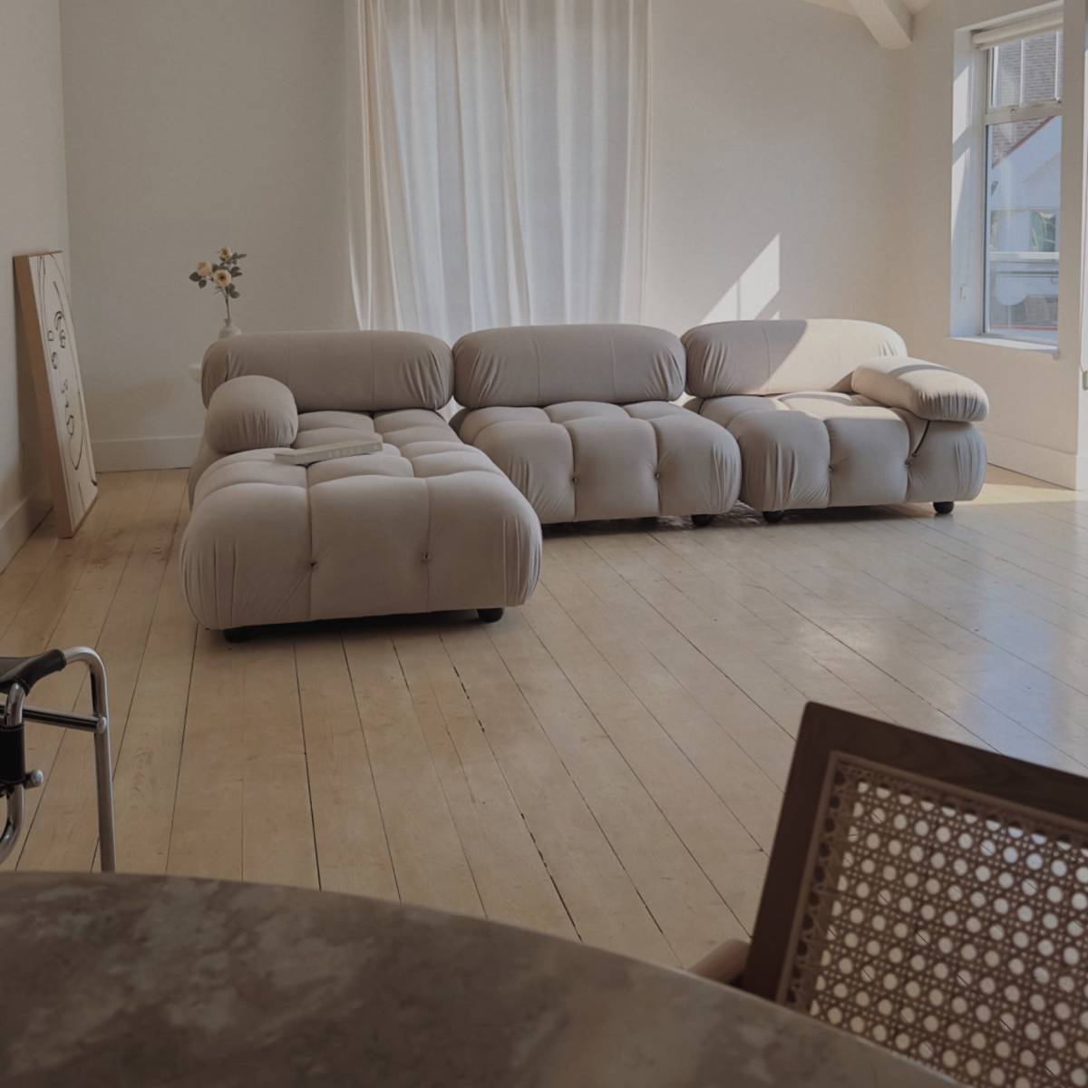 A light-colored living room with a curved beige sofa, a large window with sheer curtains, and wooden flooring.
